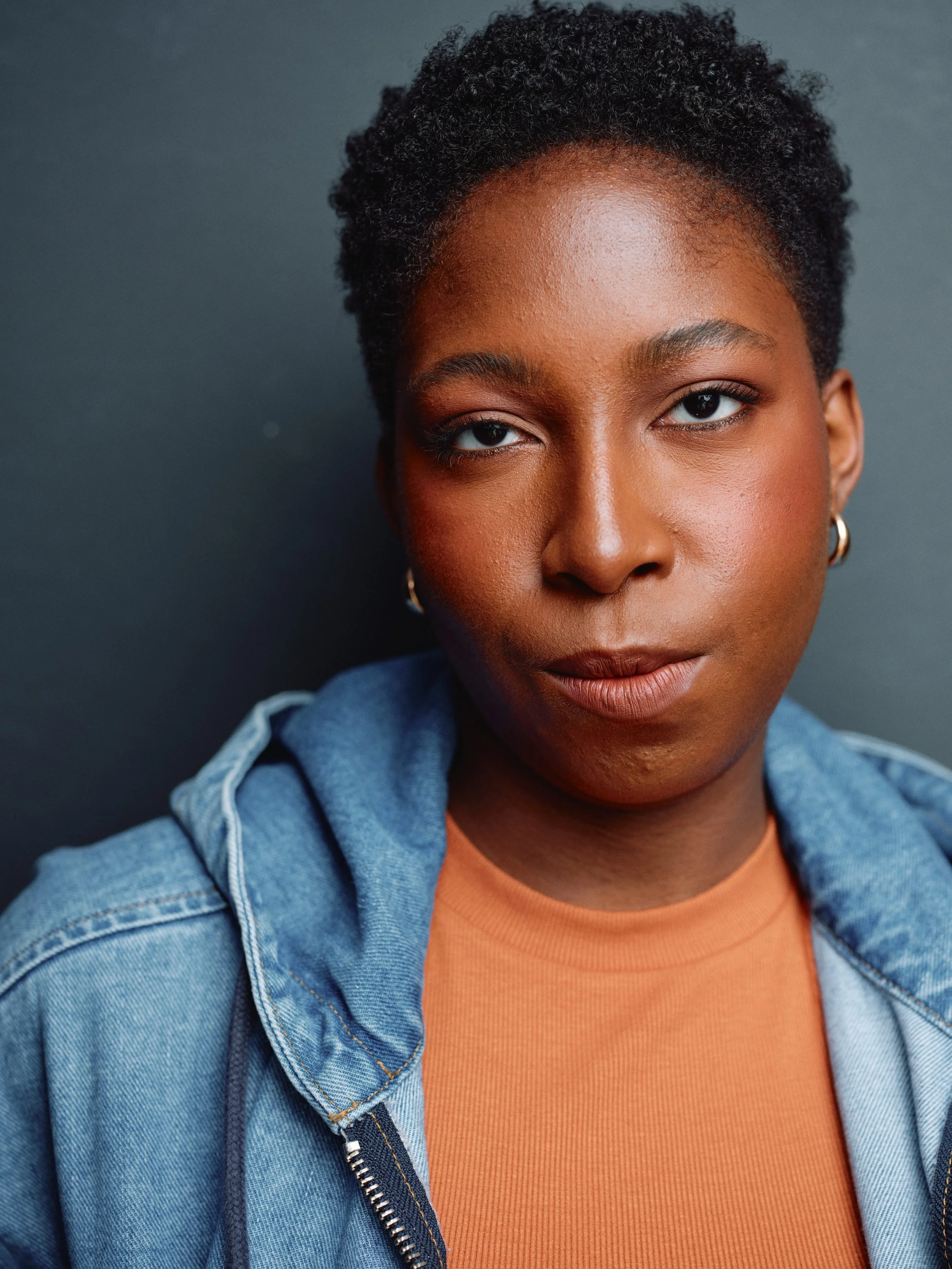 Close-up portrait of a woman with short curly hair wearing a denim jacket and an orange shirt, against a dark background.