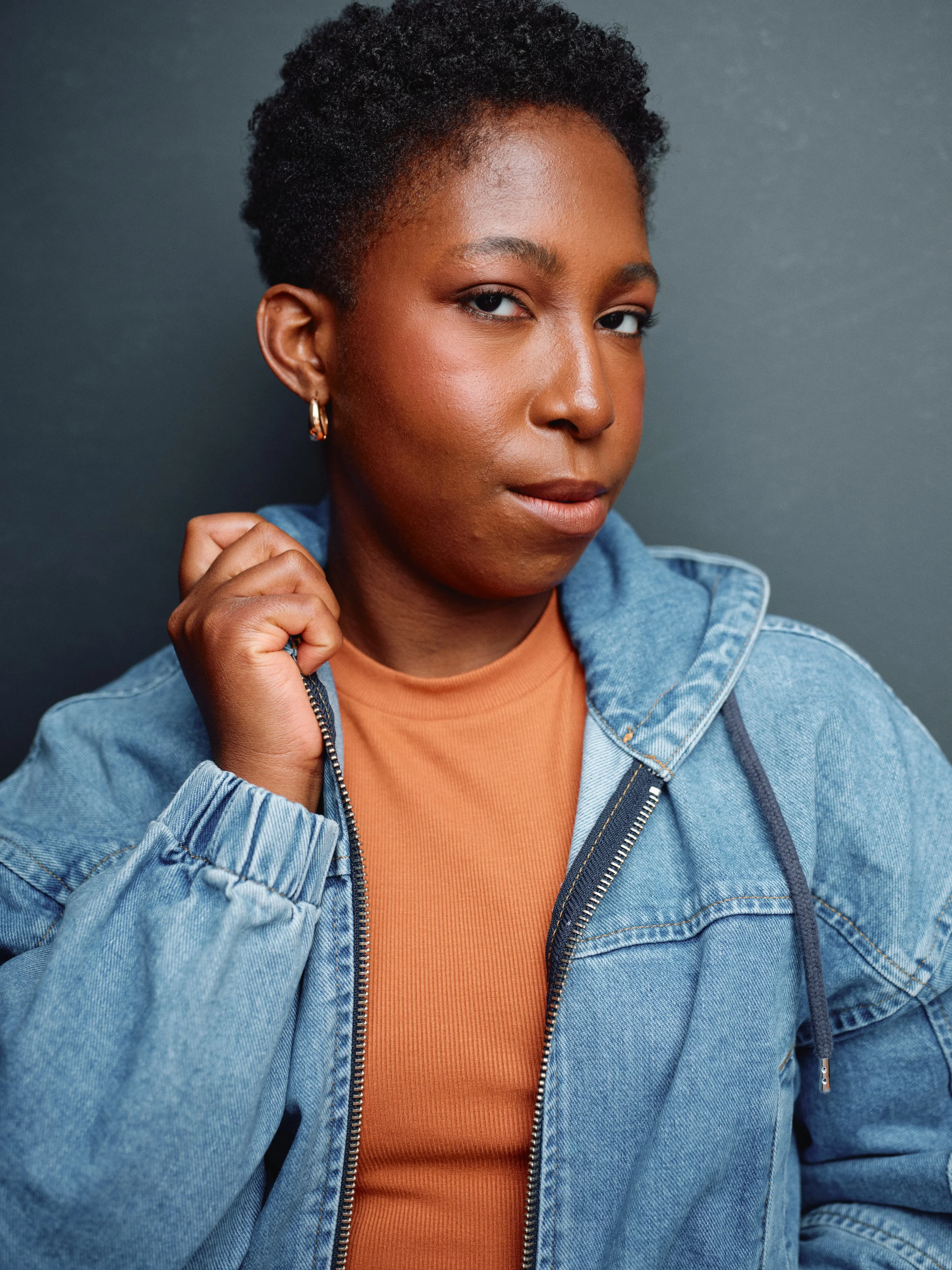 Portrait of a woman with dark skin, short curly hair, golden earrings, wearing a denim jacket over an orange shirt, standing against a dark background.