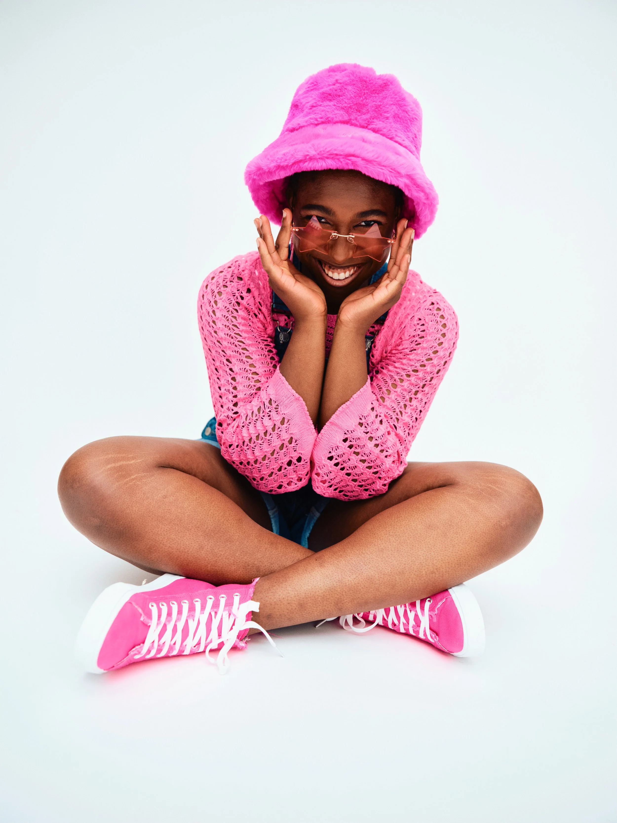 A young woman sitting cross-legged on a white background, smiling, wearing a pink fuzzy hat, pink sunglasses, a pink crochet sweater, pink sneakers, and denim shorts.