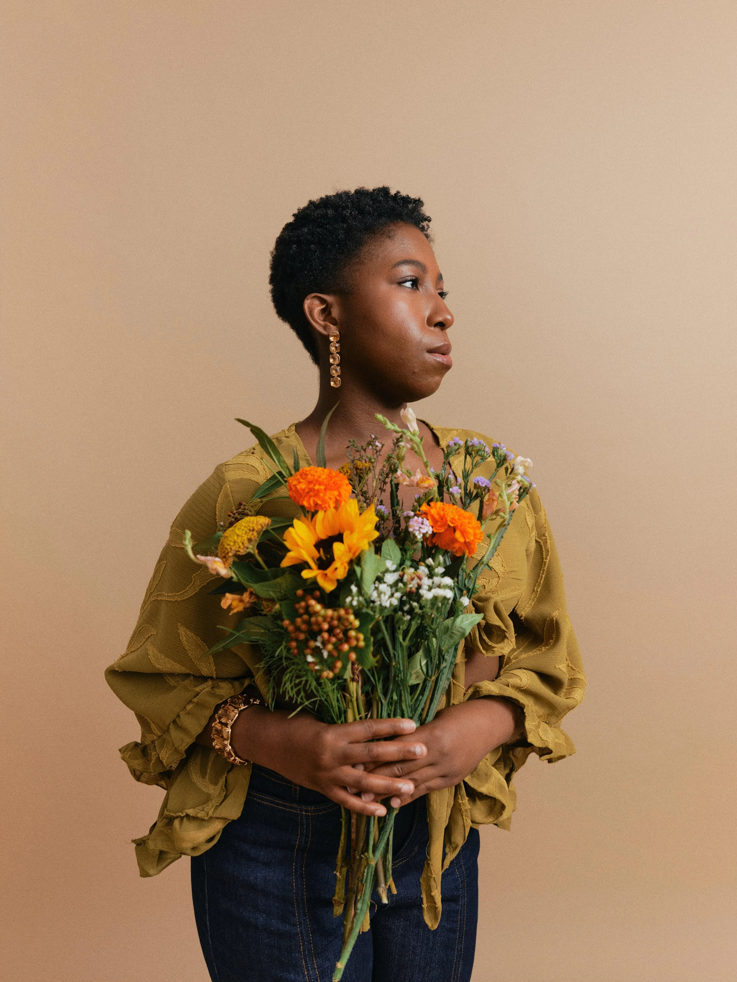 A woman with short curly hair wearing a yellow blouse and jeans, holding a bouquet of colorful flowers against a neutral background.