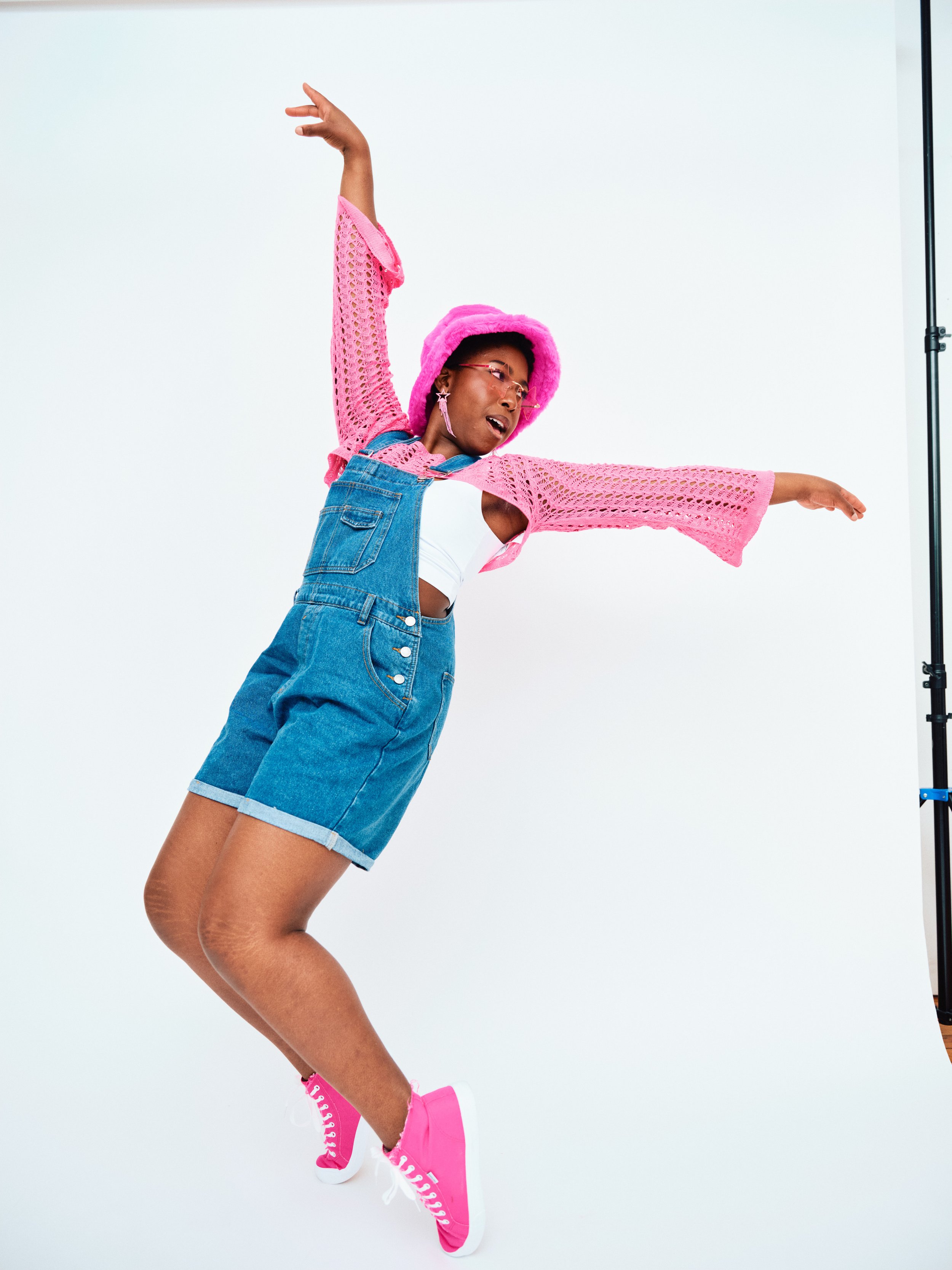 A woman dancing wearing a pink bucket hat, pink crochet jacket, white crop top, denim shorts, and pink sneakers, against a plain white background.