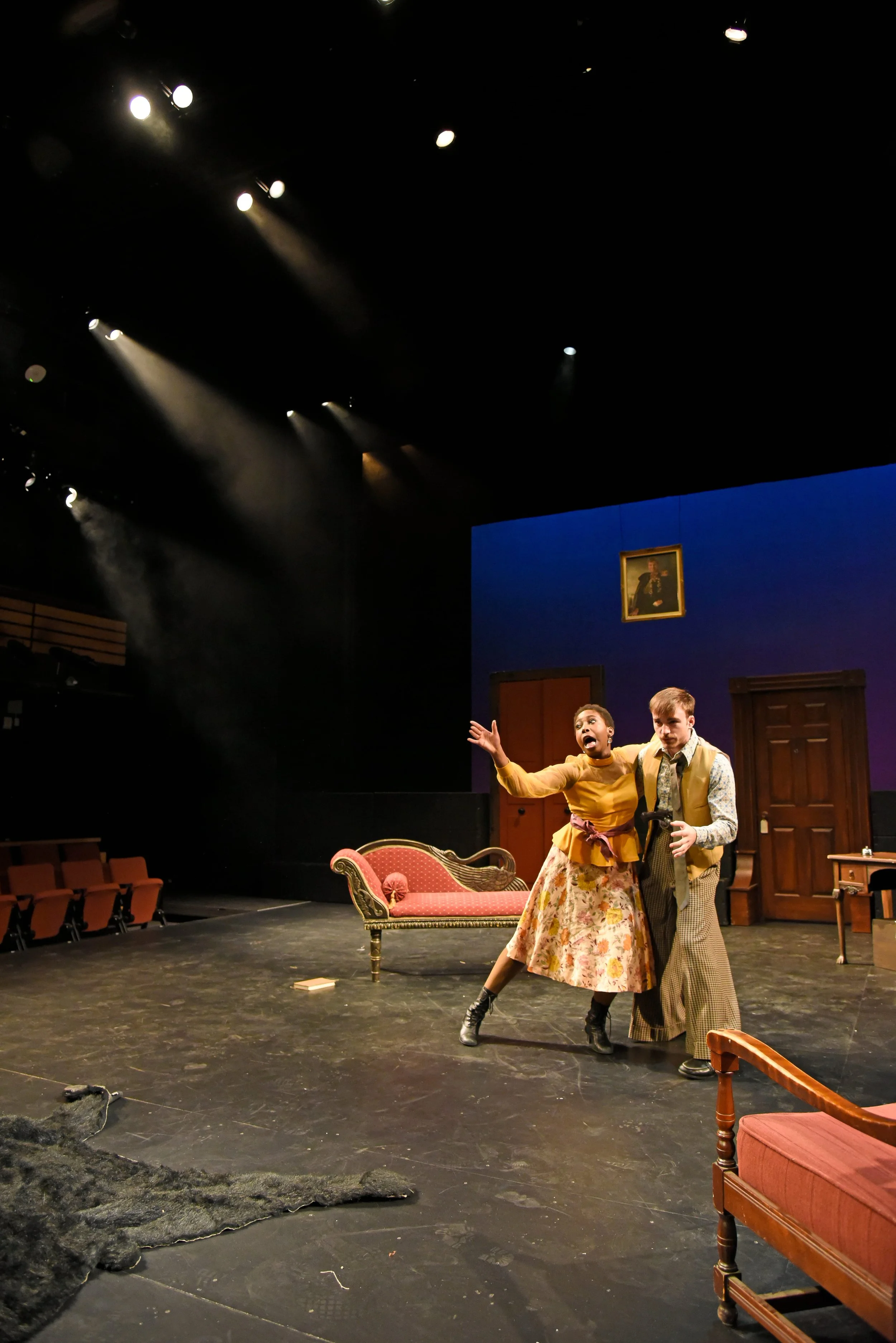 Two actors on stage in a theatrical play, surrounded by furniture, with stage lights shining down from above.
