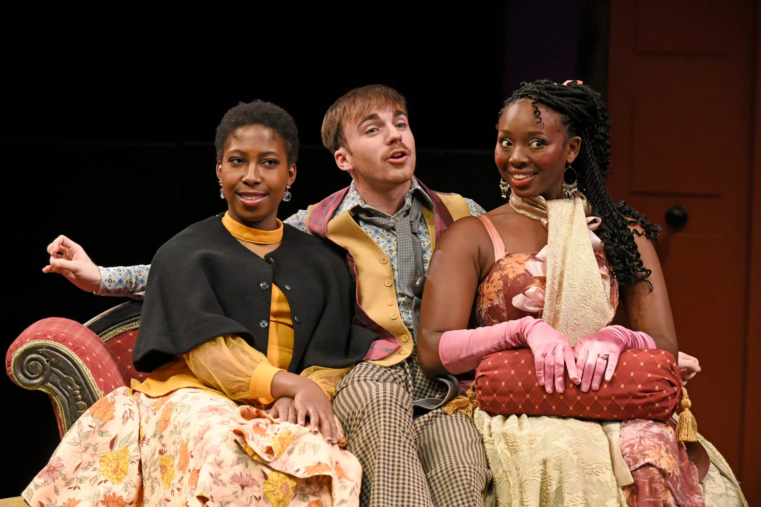 Three actors on stage, dressed in colorful vintage costumes, sitting on a ornate sofa, performing in a play.
