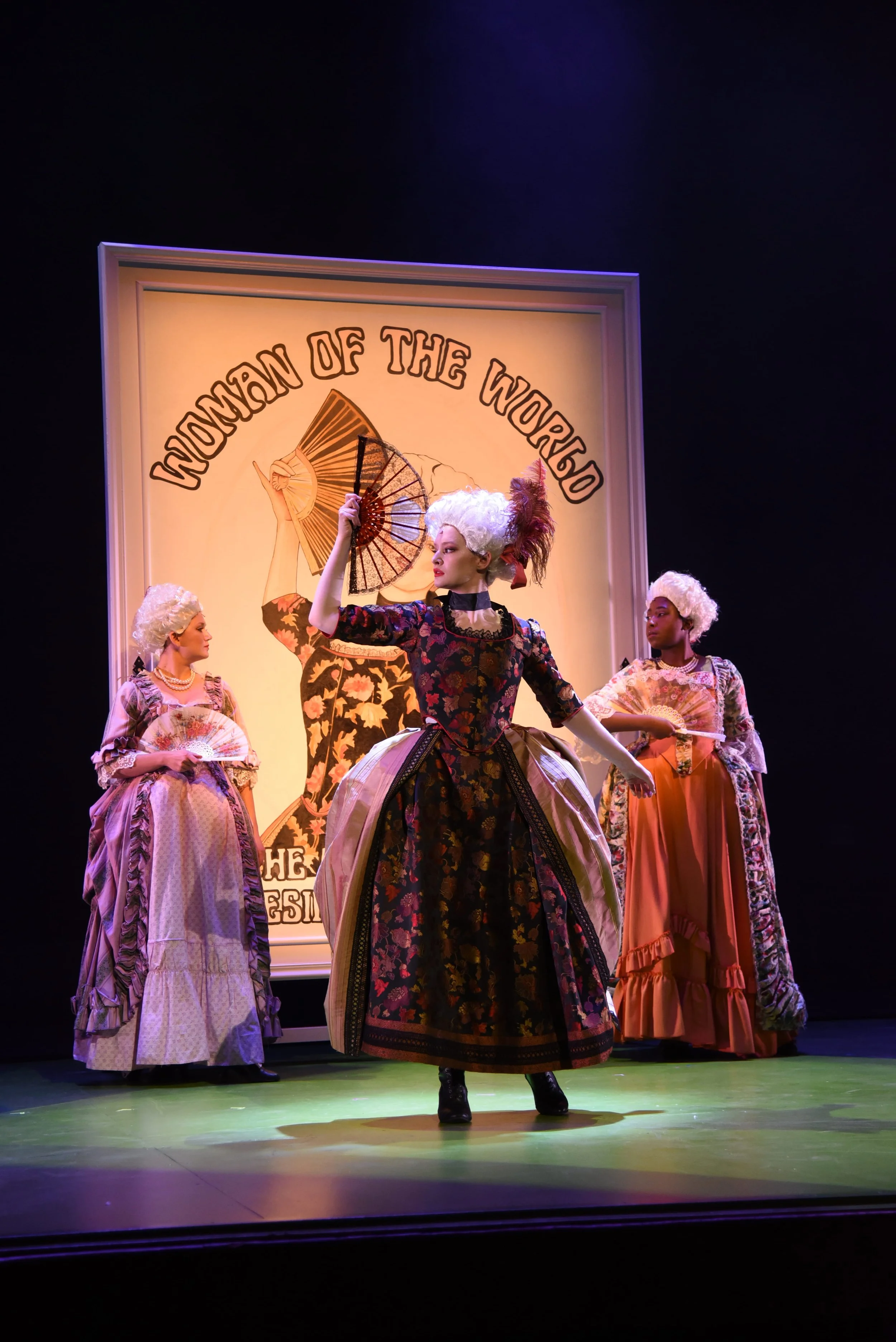Stage scene with four women in 18th-century costumes, holding fans, during a theatrical performance with a backdrop that reads 'Women of the World' and features a woman holding a fan.