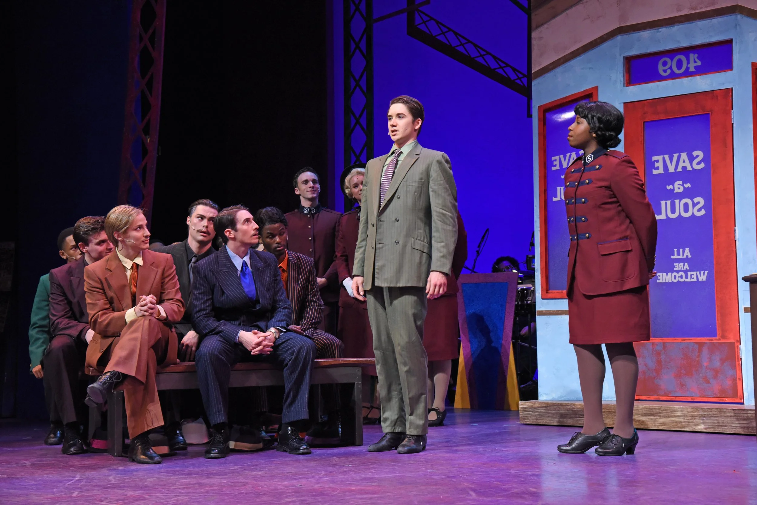 The scene is from a theatrical play featuring a group of actors dressed in vintage clothing. One man stands in front of a seated group, appearing to be speaking or performing, while a woman in a red uniform stands nearby. The setting suggests a courtroom or an official environment, marked by a backdrop with the words 'SAVE and SOUL' and a sign that reads 'AL' and 'WELCOME'.
