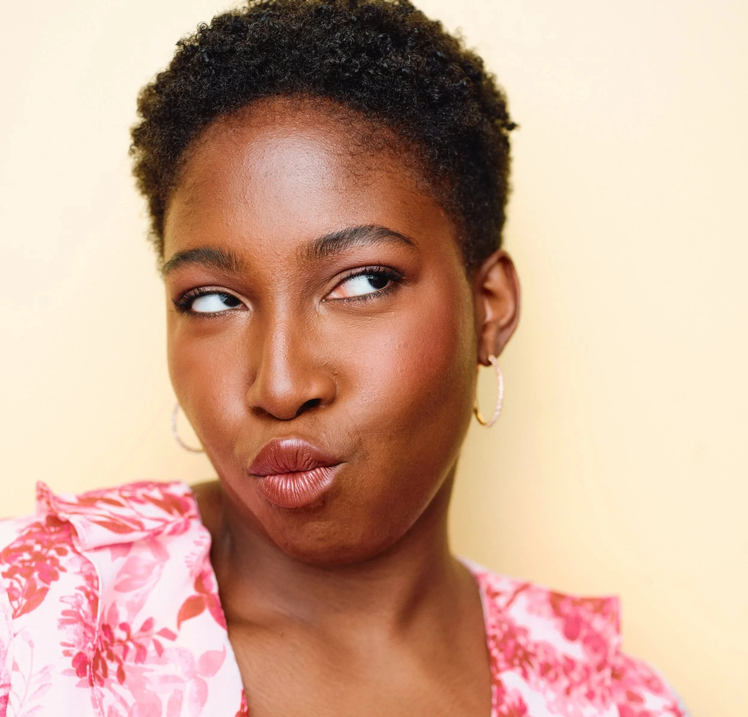 A woman with short curly hair and hoop earrings making a facial expression with her eyes rolling and lips puckered, wearing a pink floral top against a light yellow background.