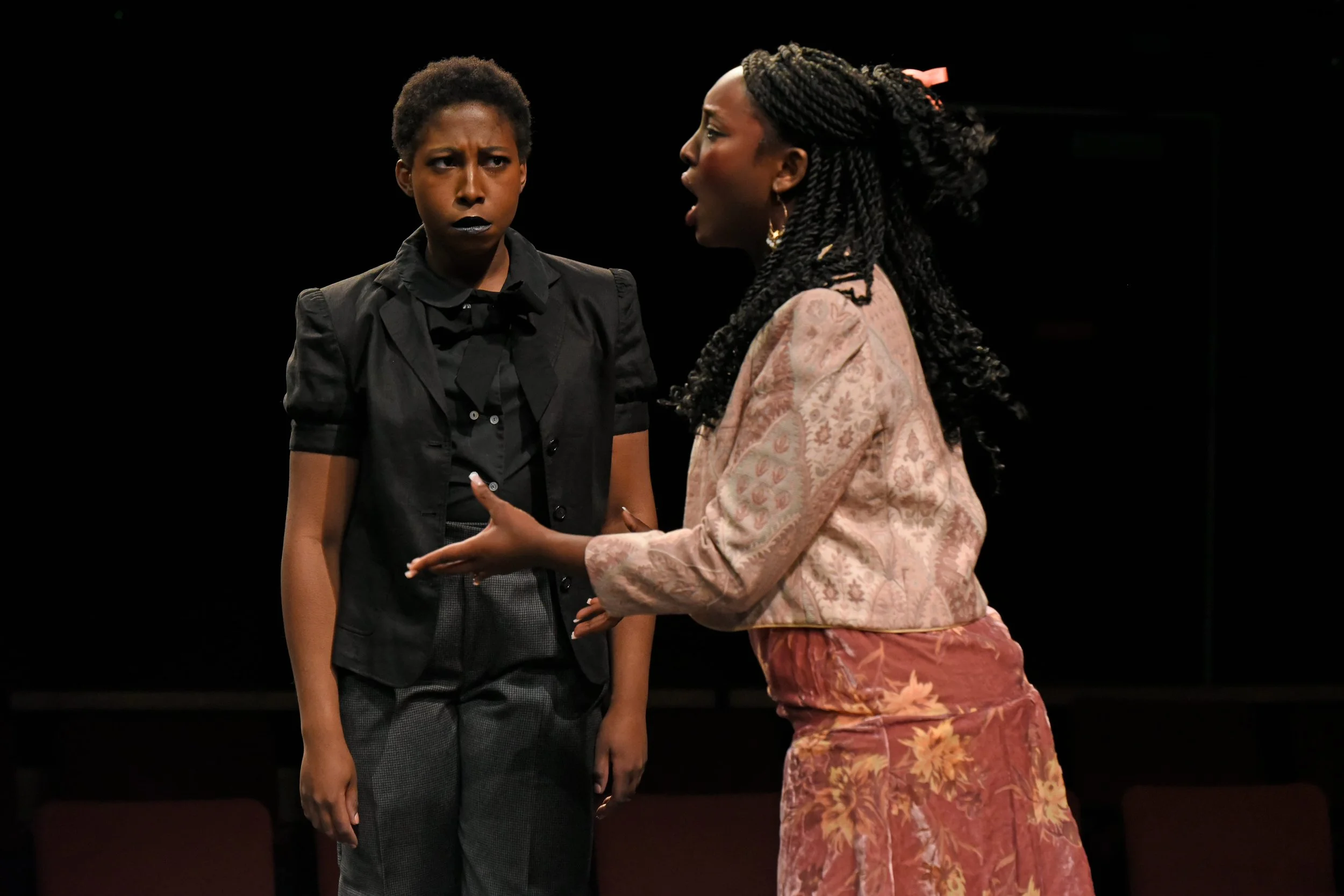 Two women engaging in a dramatic conversation on stage; one appears angry or upset, and the other looks defensive or explaining, with a dark background.