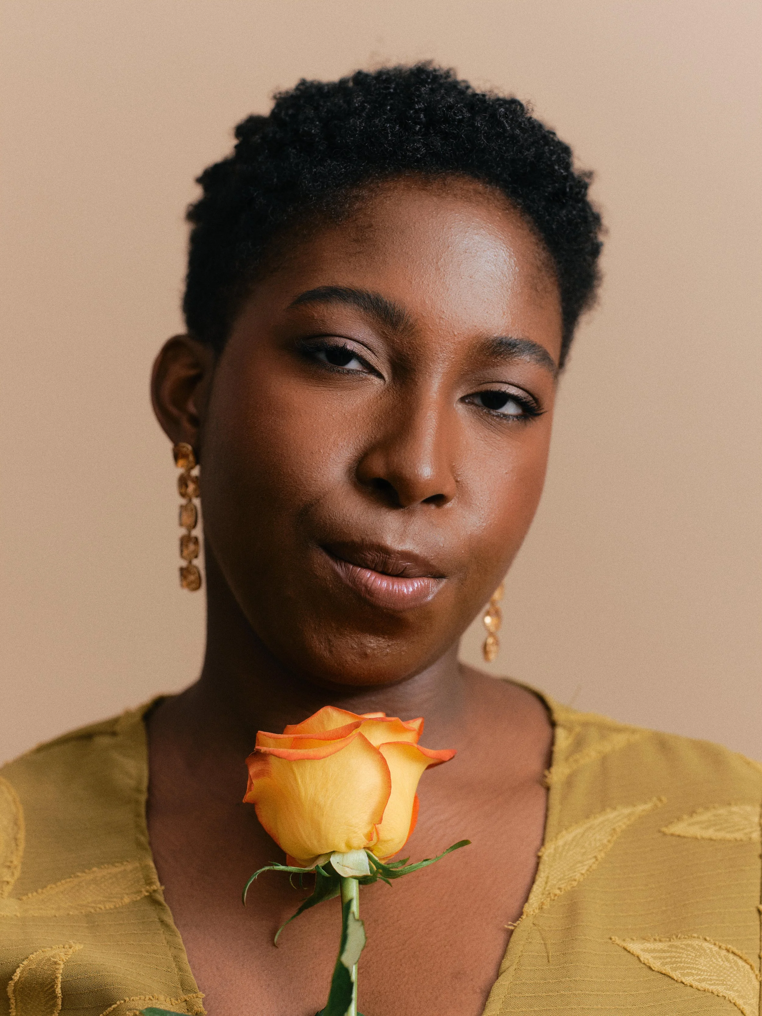 A woman with short curly hair wearing gold earrings and a yellow blouse holds a yellow and orange rose against a beige background.