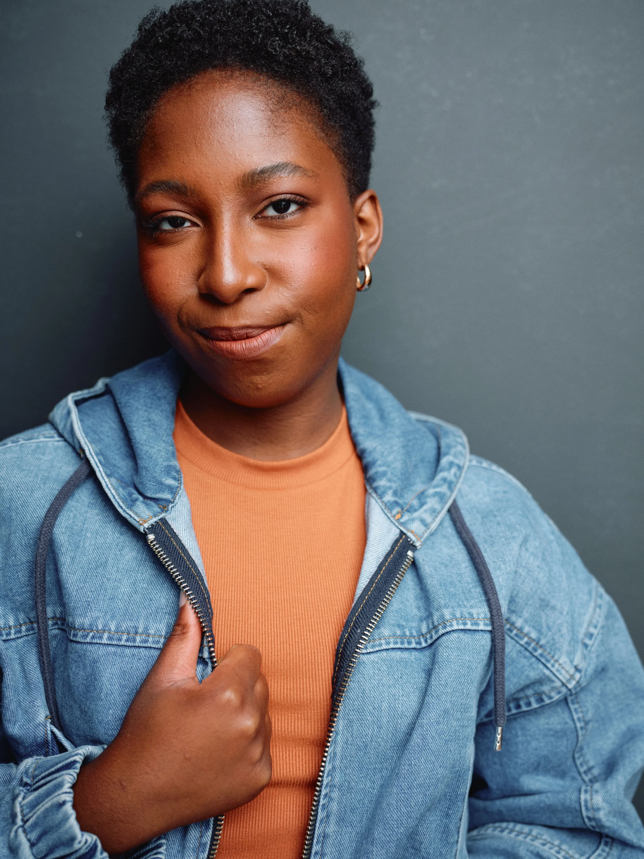 Portrait of a young African American woman with short, curly hair, wearing hoop earrings, a denim jacket over an orange shirt, and giving a thumbs-up gesture against a dark background.