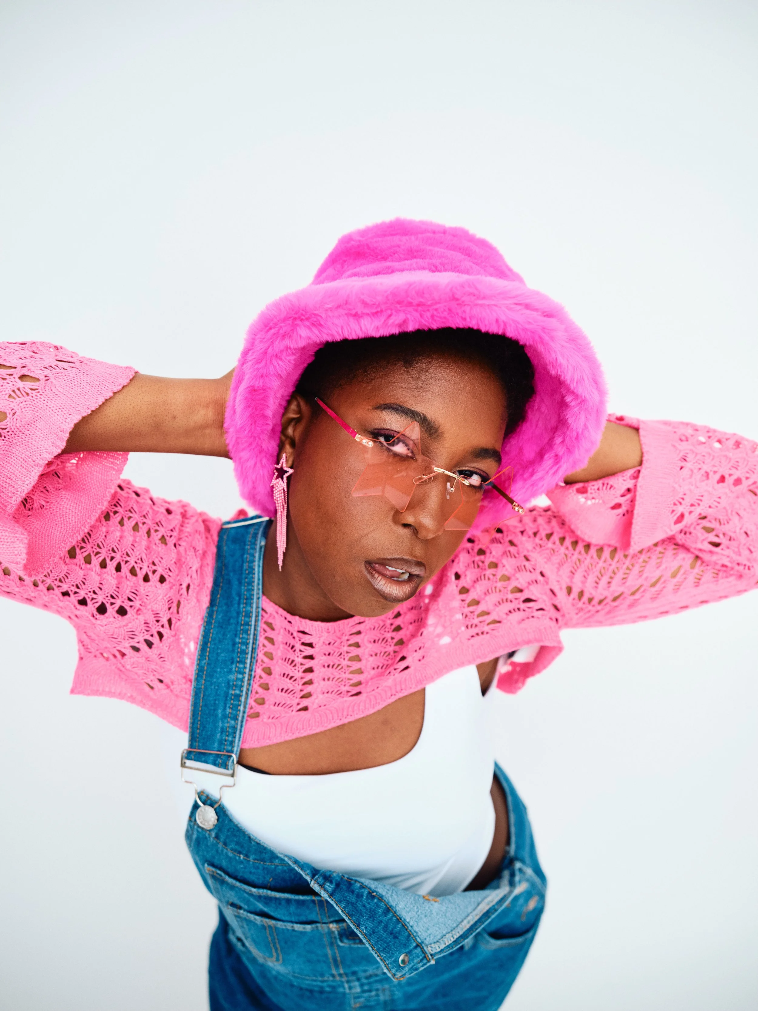 A woman with dark skin wearing pink fuzzy hat, pink sunglasses with geometric frames, pink crochet top, white tank top underneath, and blue denim overalls, posing with hands behind her head against a white background.
