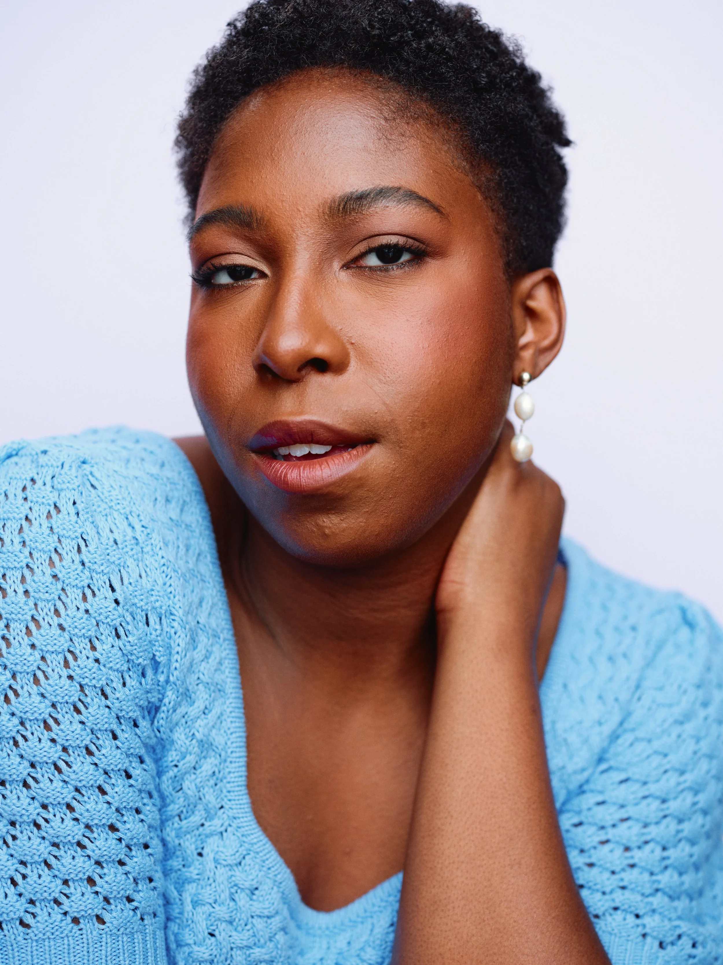 Portrait of a woman with short curly hair wearing a blue knitted top and pearl earrings, posing with one hand on her neck