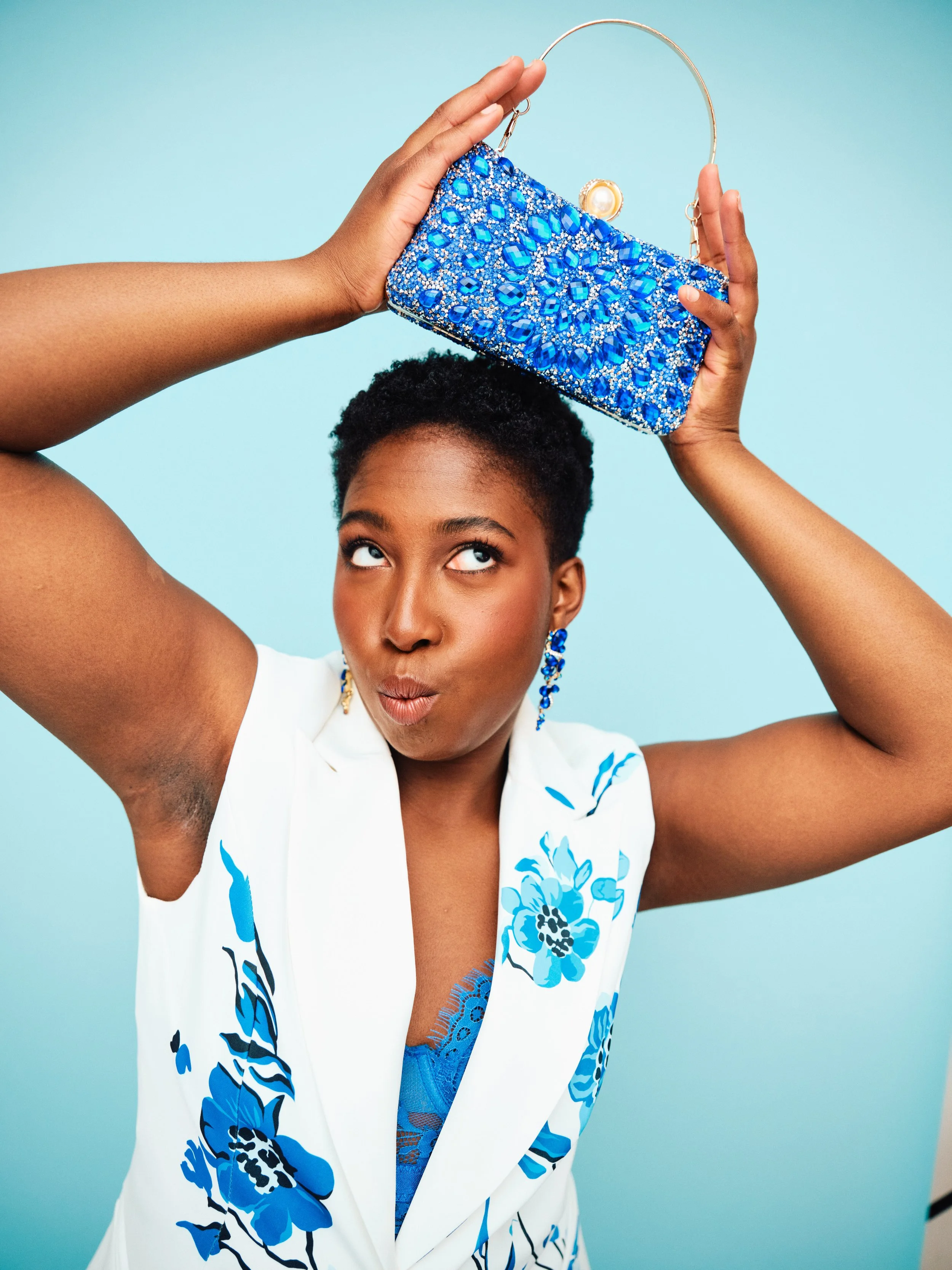 A woman with short, curly black hair and dark skin, wearing a white sleeveless dress with blue floral patterns, holds a blue beaded clutch with a pearl handle above her head, looking upward with a playful expression, against a light blue background.