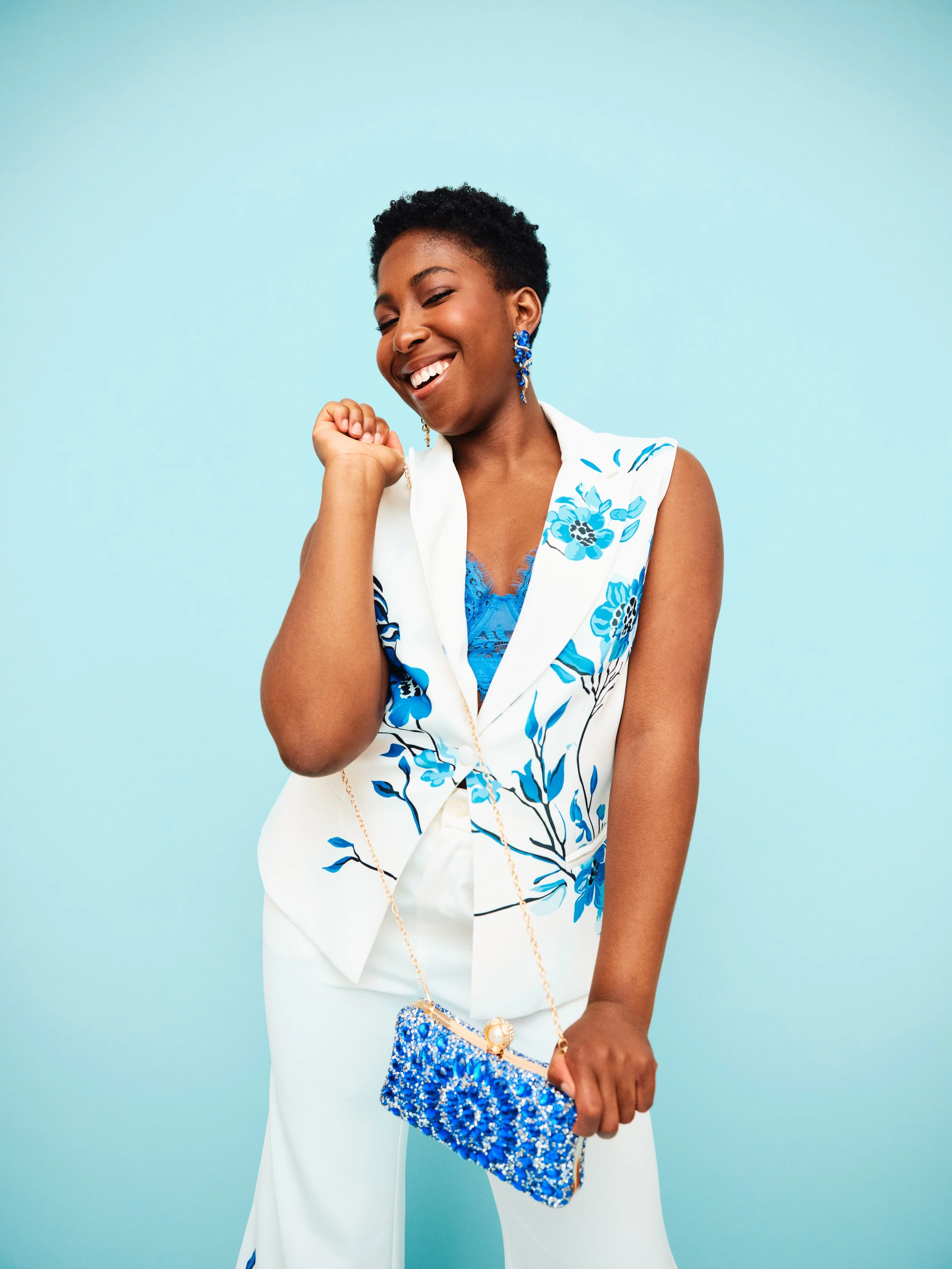 A woman dressed in white with blue floral patterns, smiling, holding a blue beaded clutch, and standing against a light blue background.