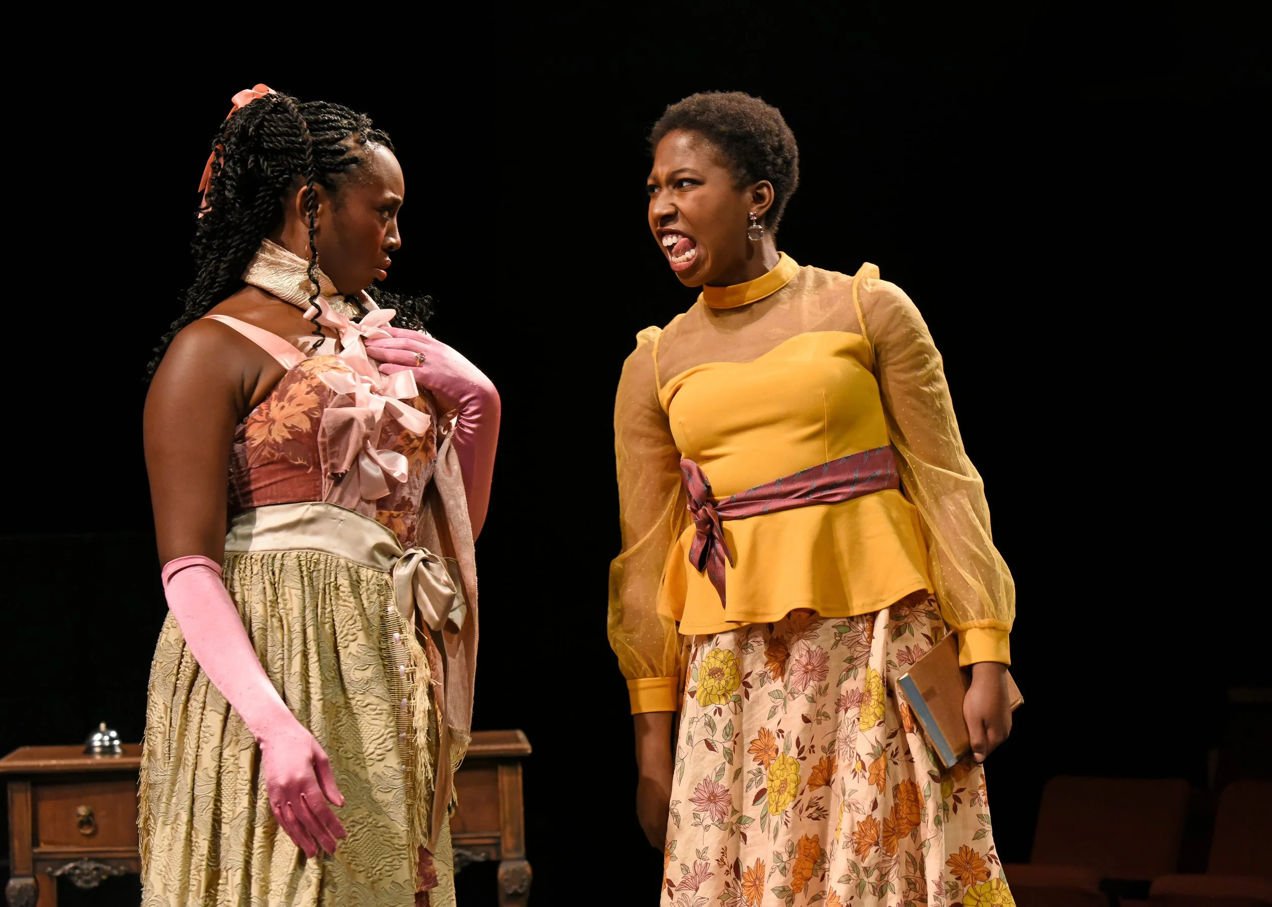 Two women on stage, one with braids and pink gloves looking down, the other with short hair and an angry expression, holding a book and wearing a yellow top and a floral skirt.