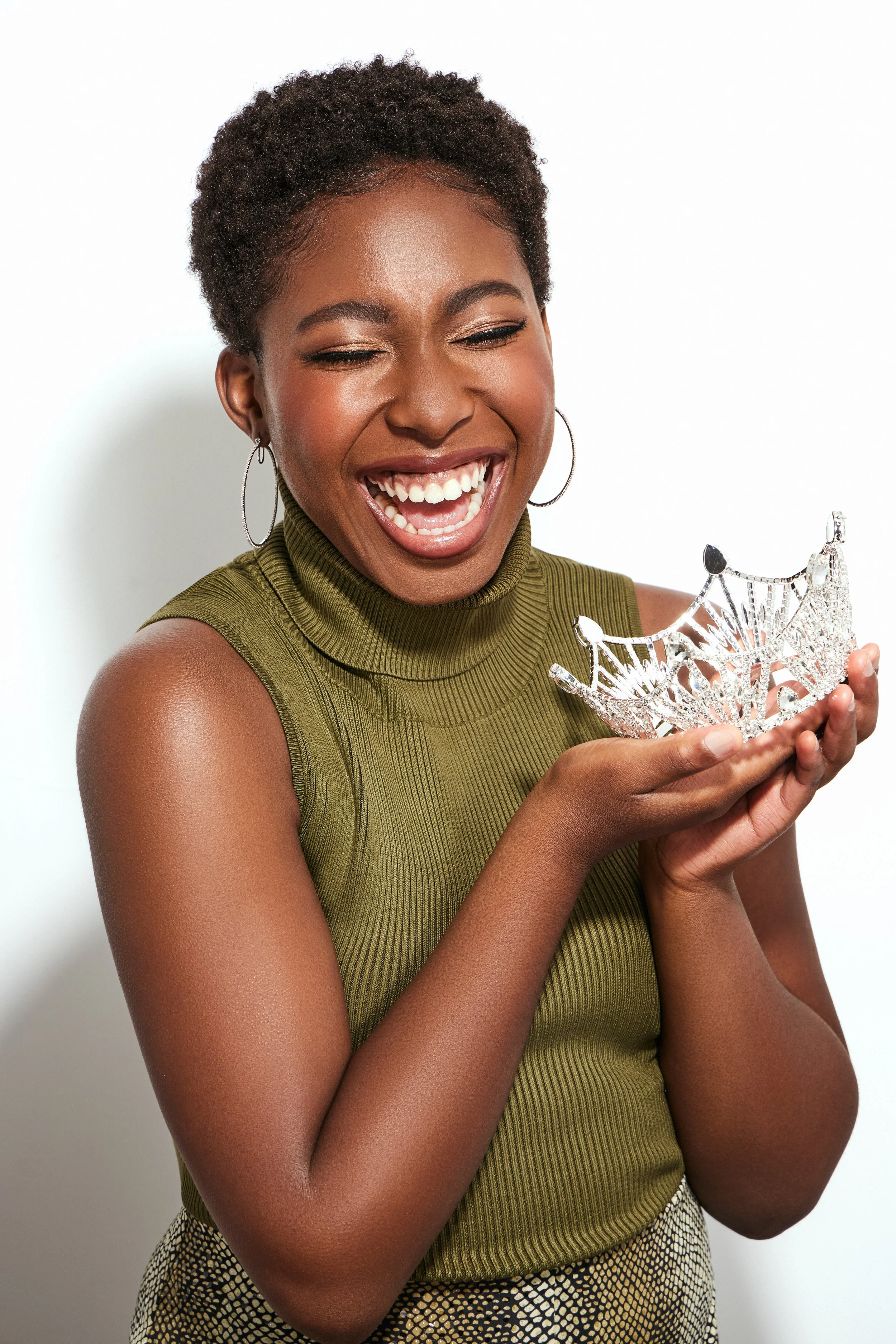 A woman with short curly hair smiling and holding a silver tiara in her hand, wearing a sleeveless green turtleneck top and hoop earrings.