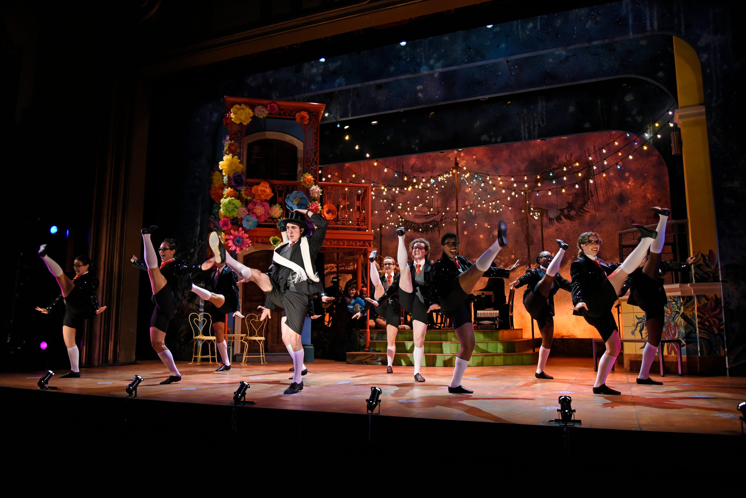 A theatrical dance performance on stage with dancers in black costumes and white socks, performing high kicks. The background features a painted scene with string lights, a decorated balcony with colorful flowers, and a whimsical, starry sky.