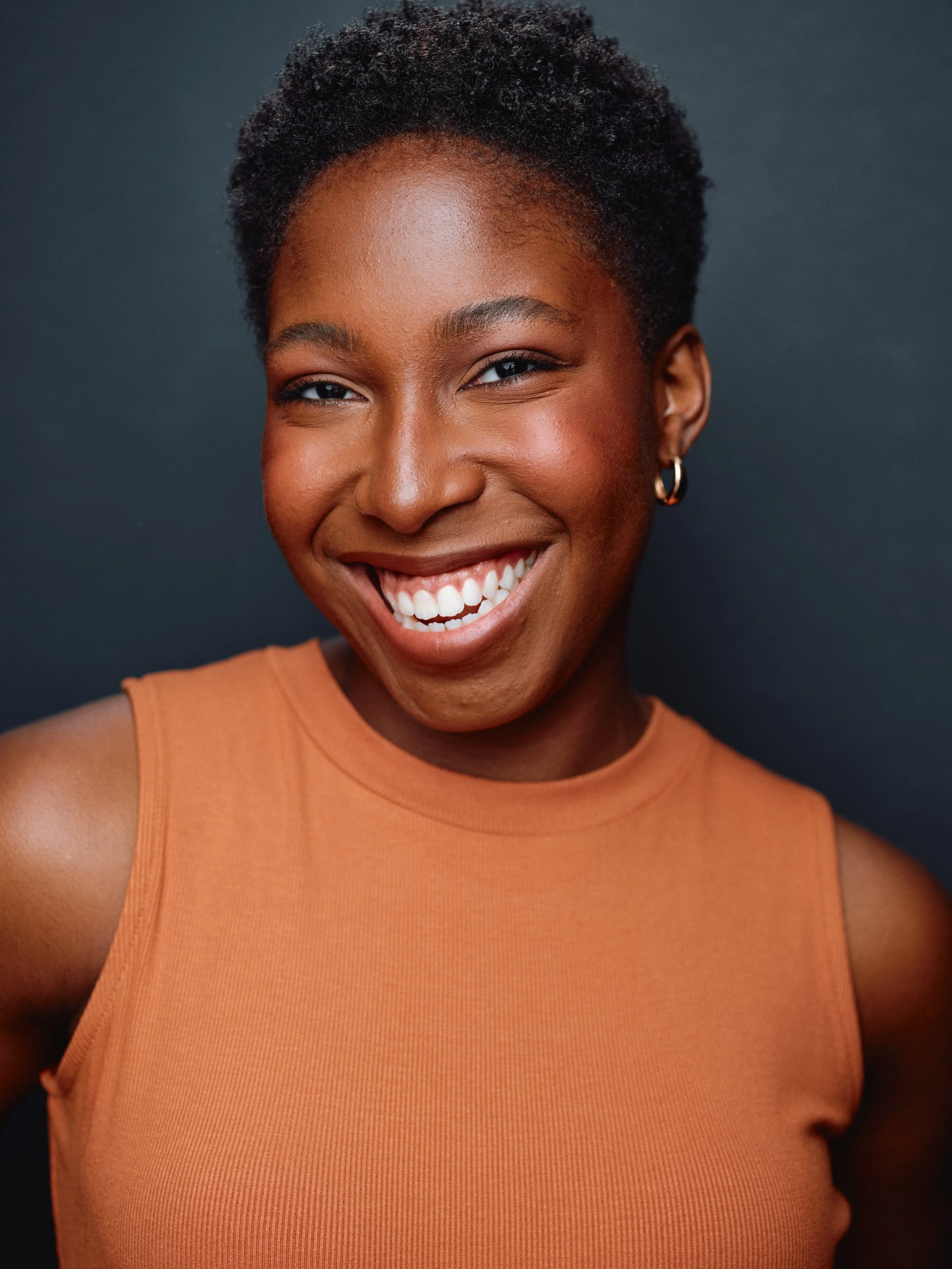 A woman with short curly black hair, wearing a sleeveless orange top and hoop earrings, smiling in front of a dark background.