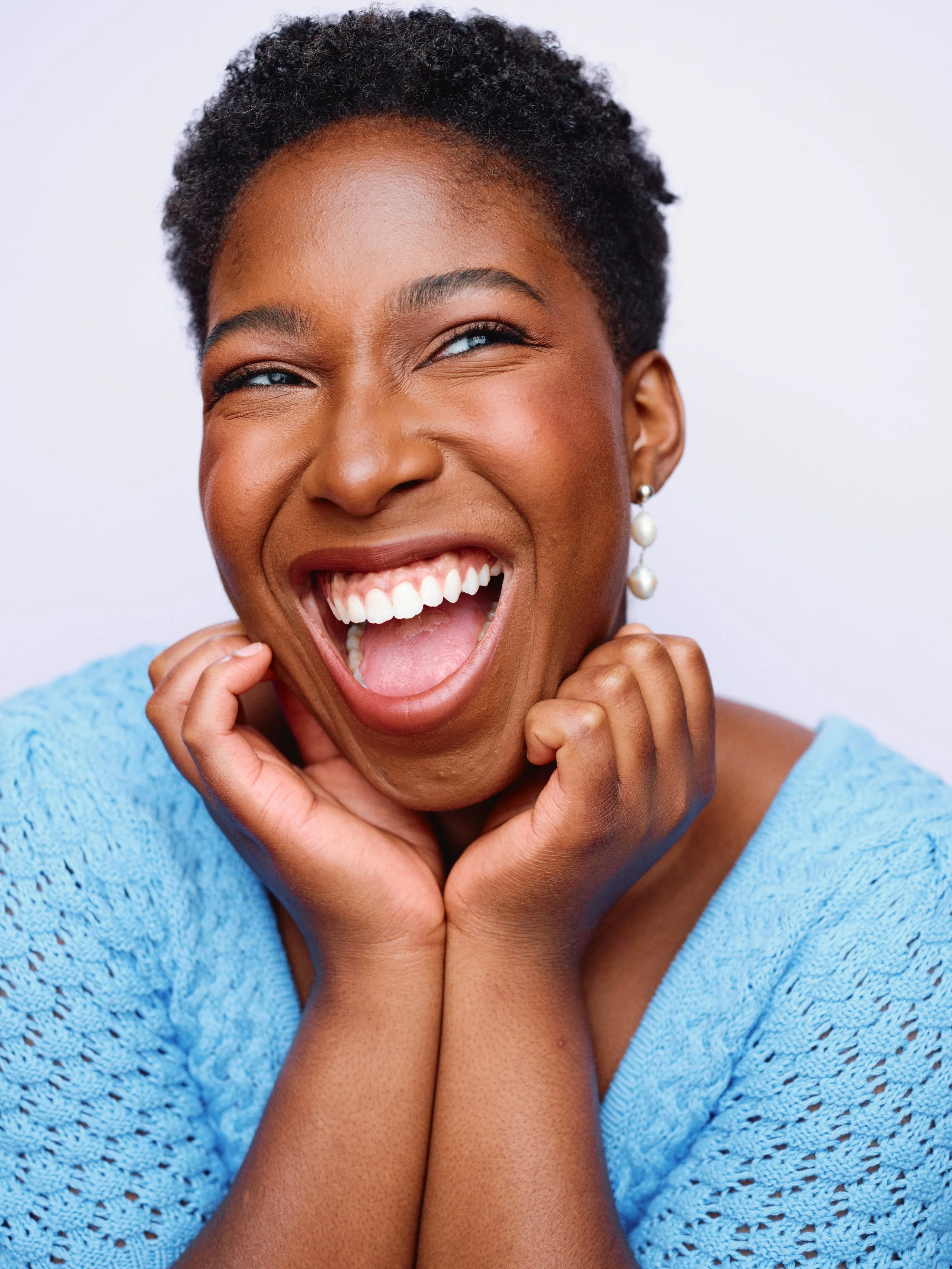 Close-up of a joyful woman with short curly hair, wearing a light blue knit top, laughing with her mouth open, hands gently holding her chin, and wearing pearl earrings against a plain white background.