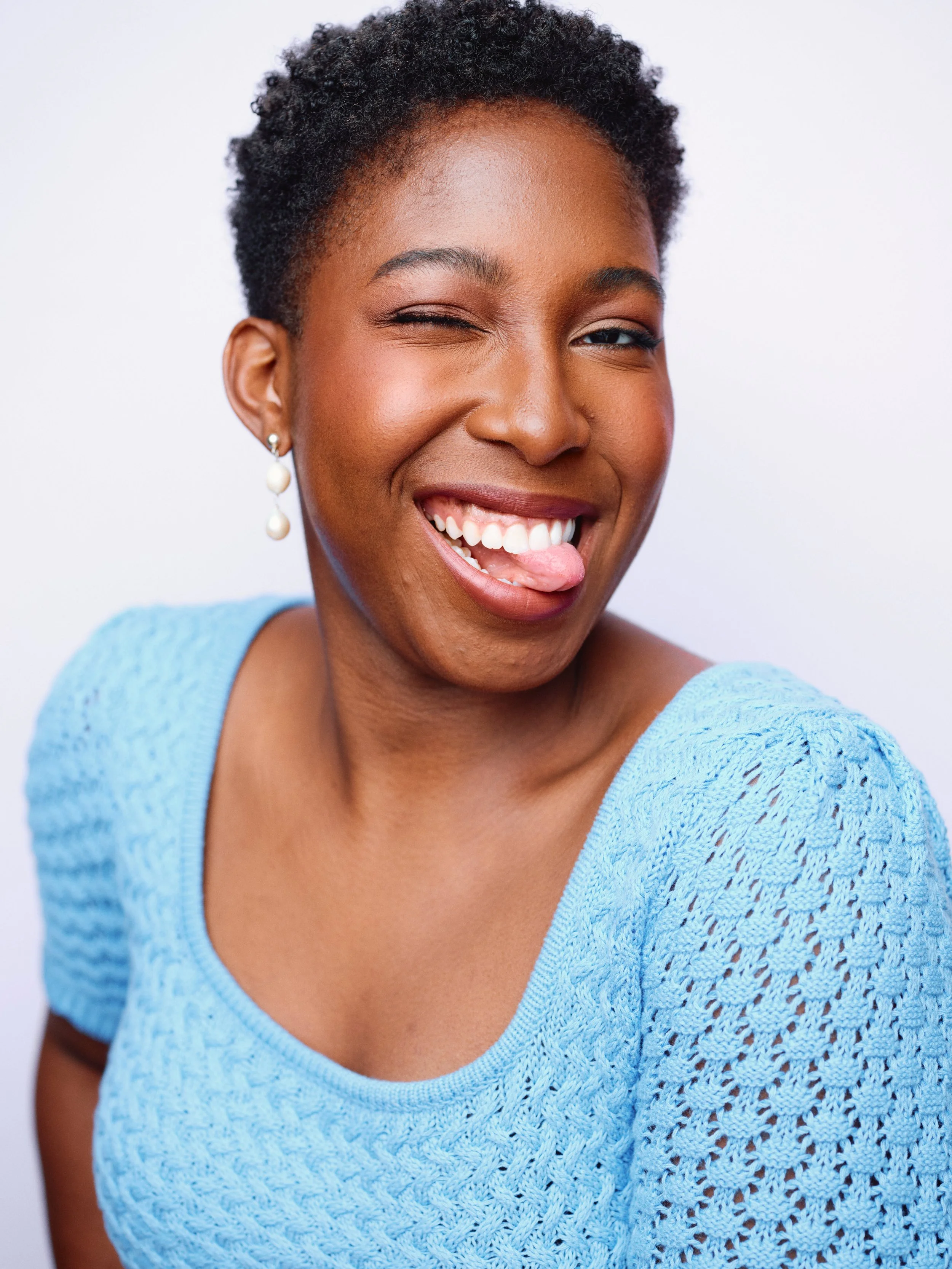 A woman with short, curly hair, wearing a blue knitted top and pearl earrings, is smiling playfully with her tongue out and winking.