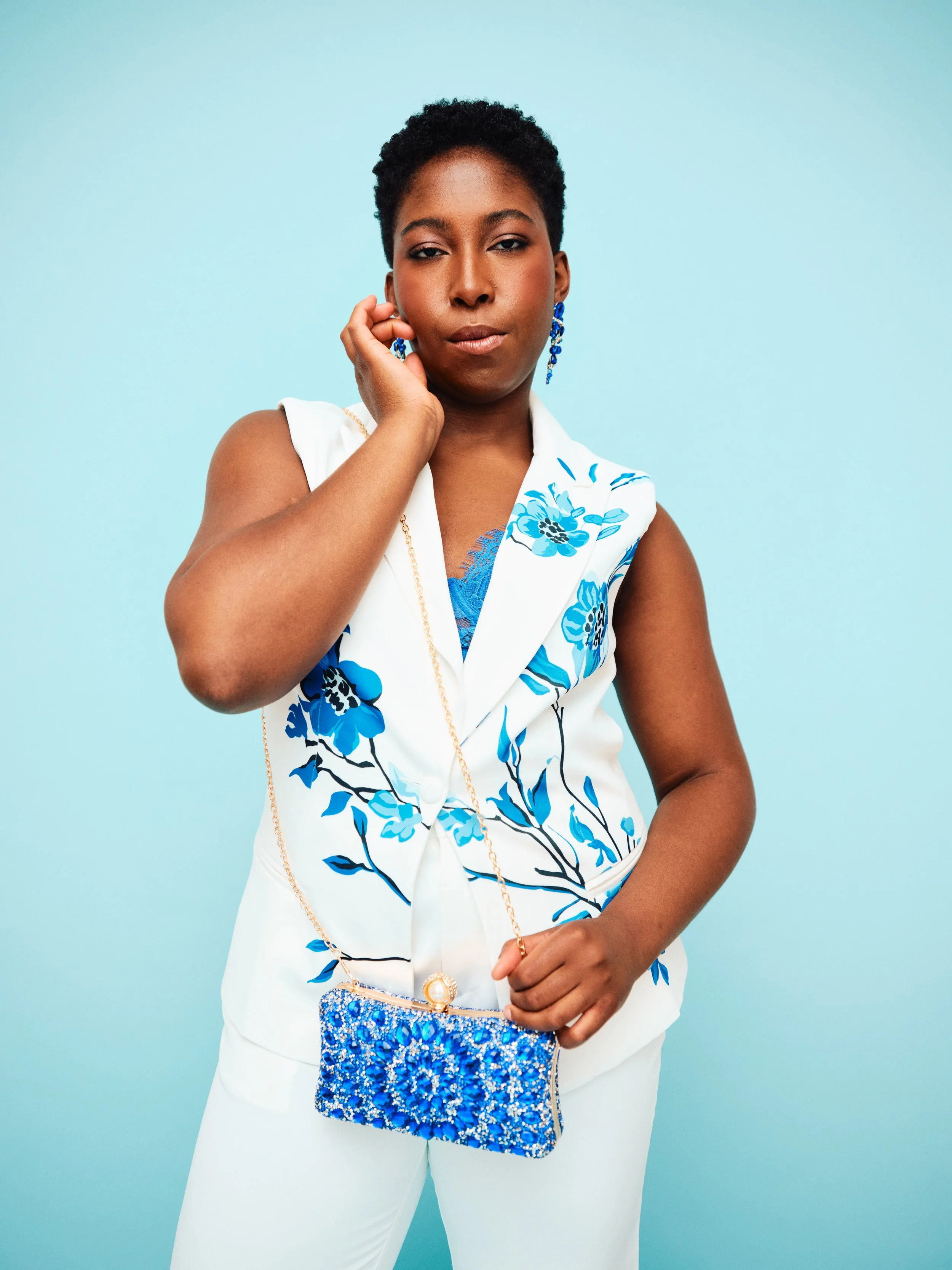 A woman with short curly hair wearing a white sleeveless blazer with blue floral pattern, white pants, and blue earrings, holding a blue beaded purse against a light blue background.