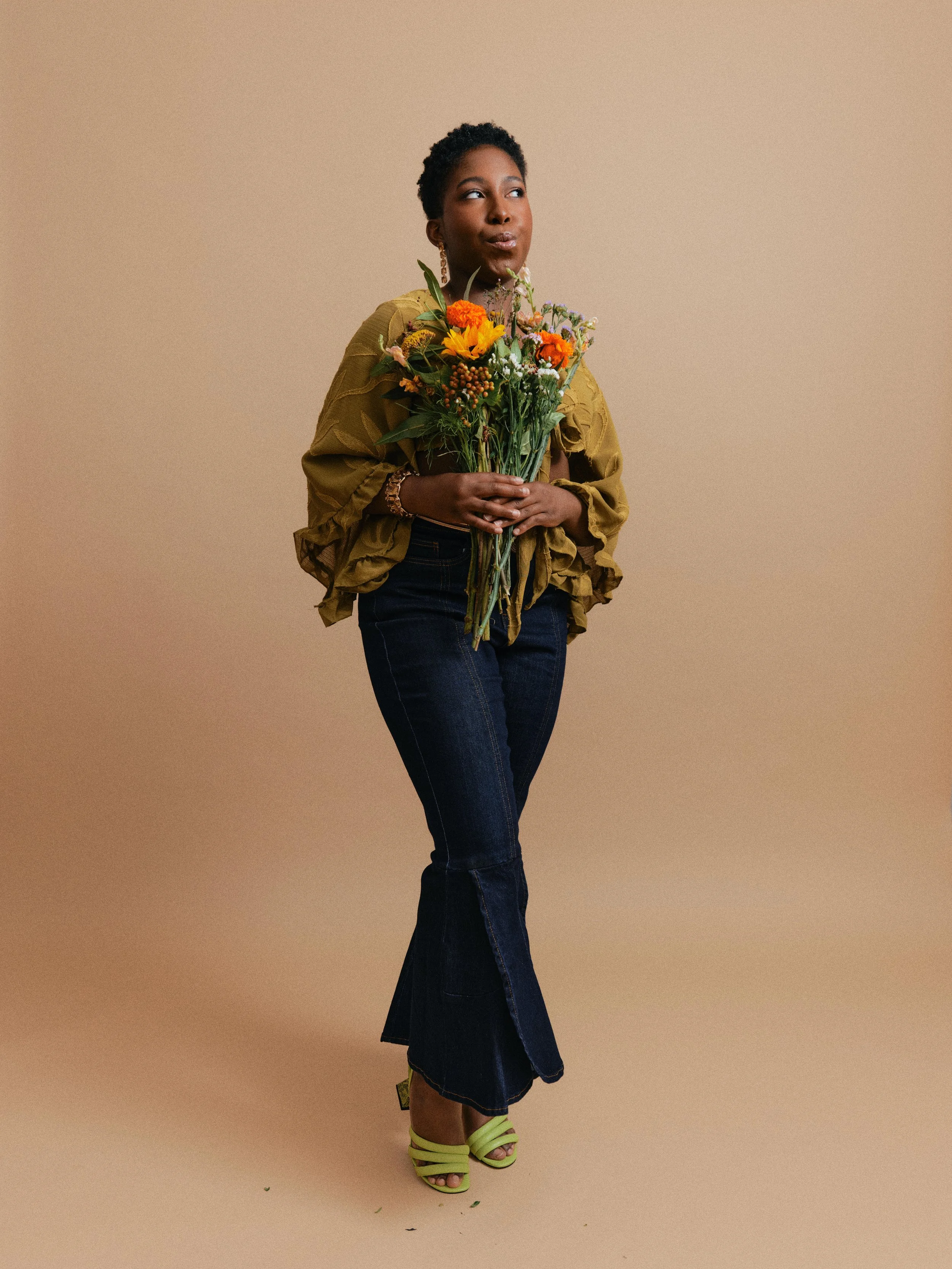 A woman with short curly hair wearing a mustard yellow blouse, dark jeans, and lime green heels holding a bouquet of orange, yellow, and white flowers against a beige background.