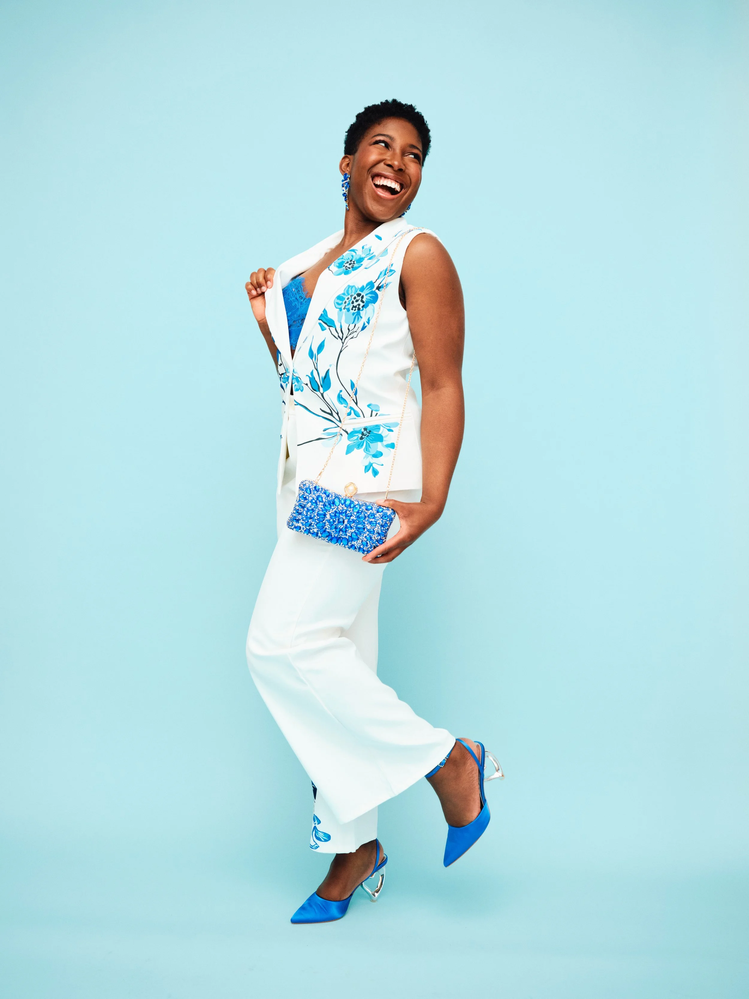 A woman smiling and posing against a light blue background, wearing a white sleeveless suit with blue floral embroidery, a blue top, matching blue earrings, blue shoes with clear heels, and holding a sparkly blue clutch bag.