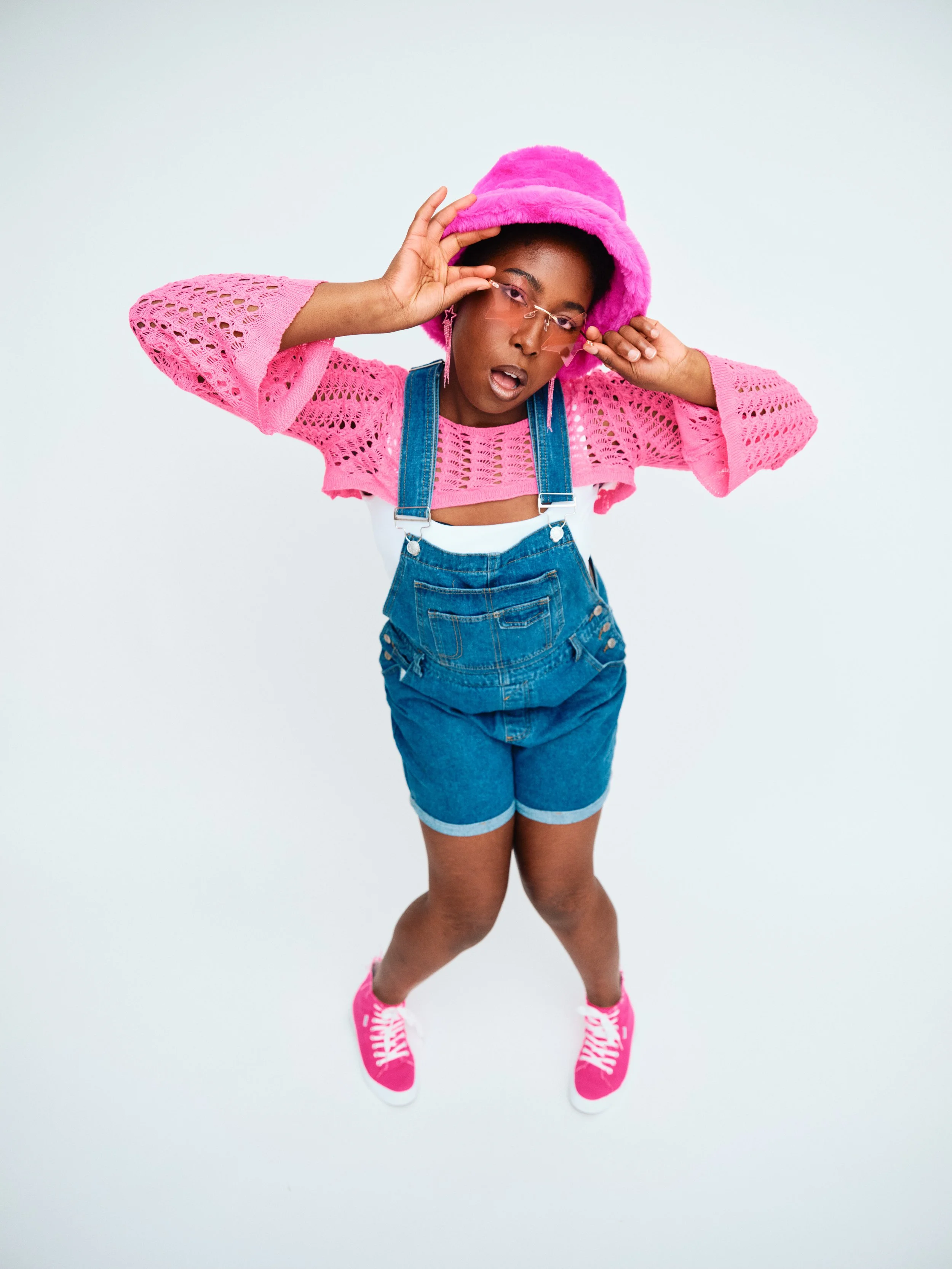 Young woman wearing a pink fuzzy bucket hat, pink crochet top, denim overalls, and pink sneakers, adjusting her pink sunglasses and looking at the camera.