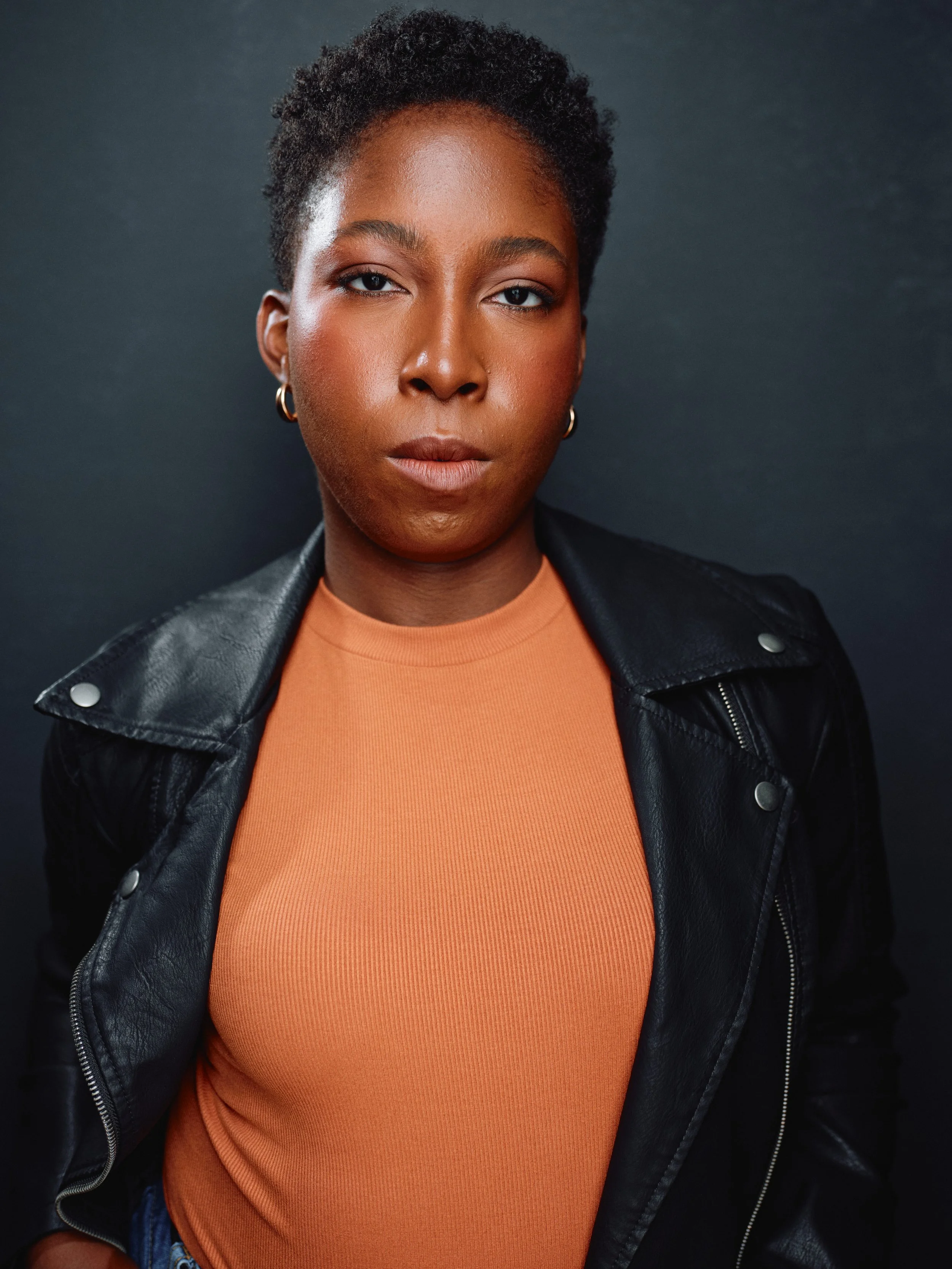 A confident woman with natural short hair, wearing a black leather jacket and an orange top, standing against a dark background.