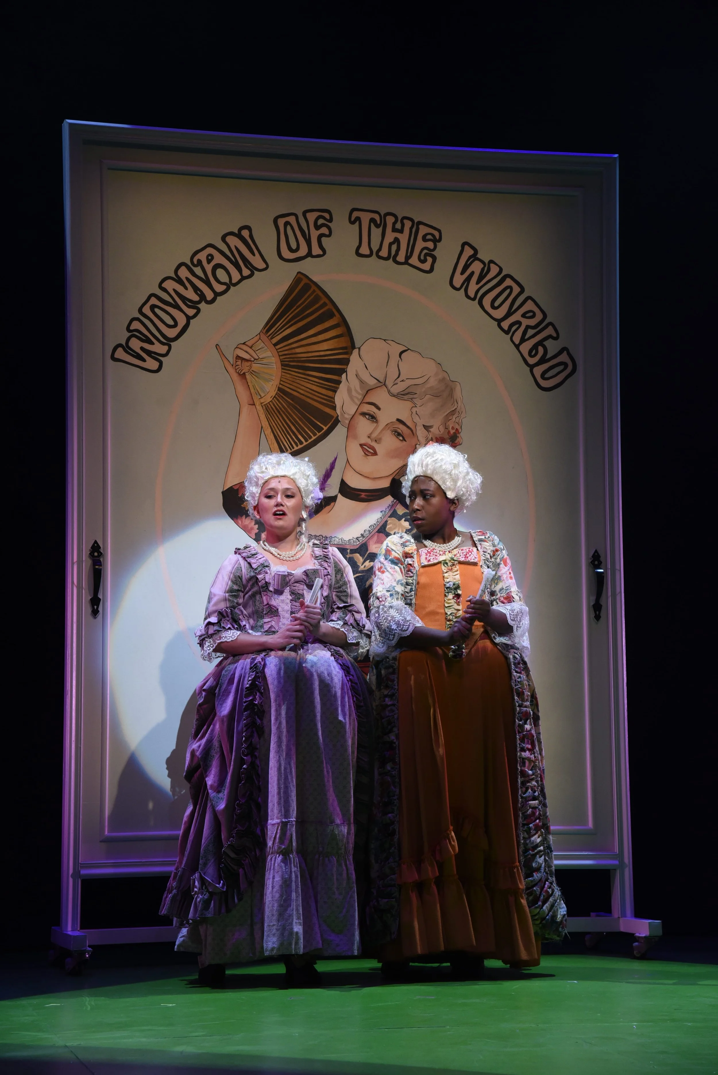 Two women in 18th-century costumes, with powdered wigs, standing on stage in front of a large sign that reads 'Women of the World' and features an illustration of a woman with a fan.