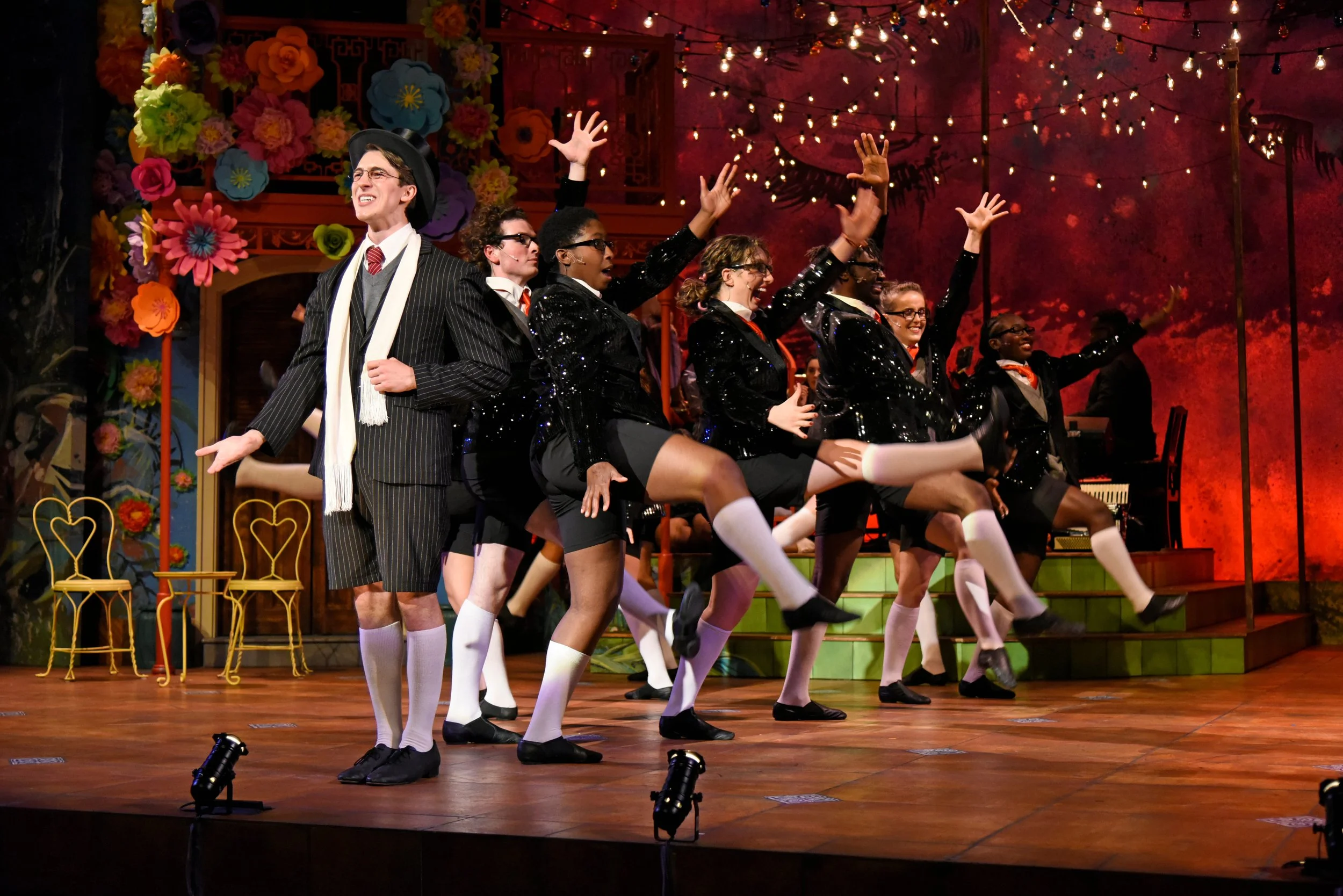 Actors performing a scene from a musical or play on a stage with festive decorations, including colorful paper flowers and string lights, with some actors in black sequined costumes and others in vintage-style suits.