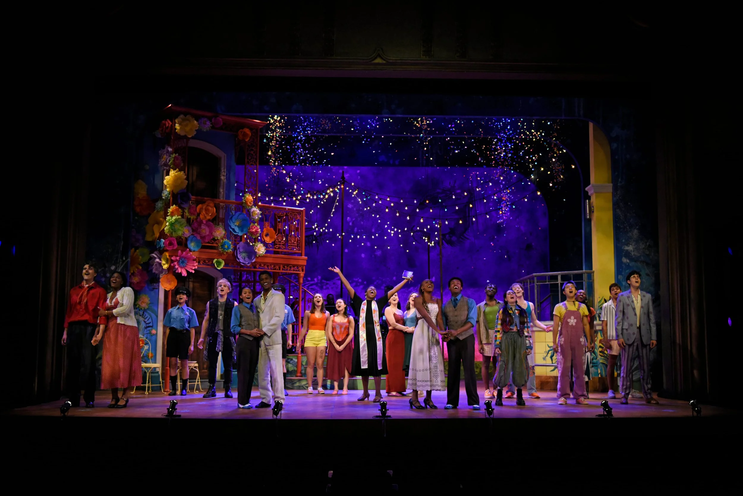 The cast of a vibrant theatrical musical on stage, with colorful costumes, floral decorations, and sparkling lights in the background, as performers take a bow.