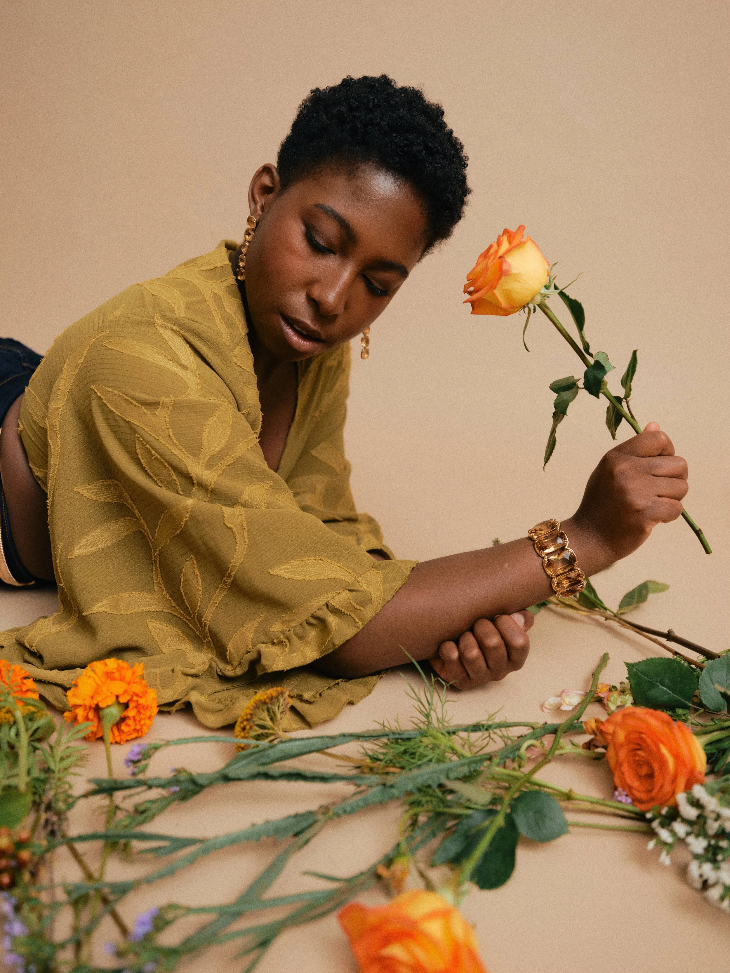 A young woman with short, curly hair lying on the floor, wearing a yellow blouse with leaf patterns. She is holding a yellow and orange rose and surrounded by various colorful flowers on a beige background.