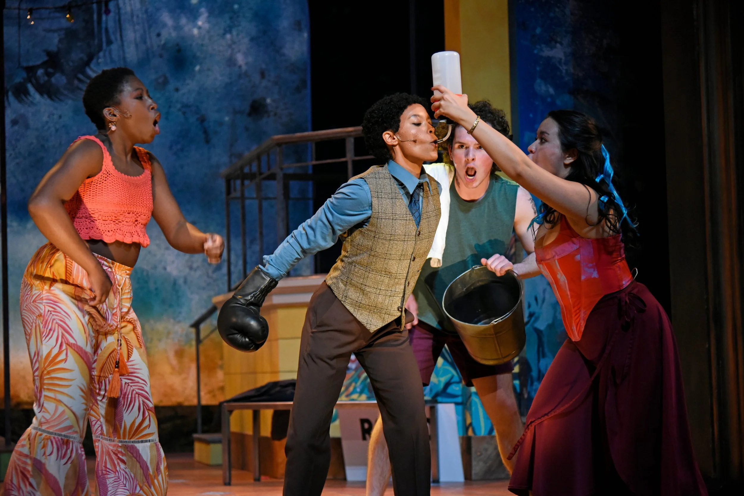 Actors on stage performing a heated scene with one woman holding a plastic bottle and a bucket, others showing expressive emotions.