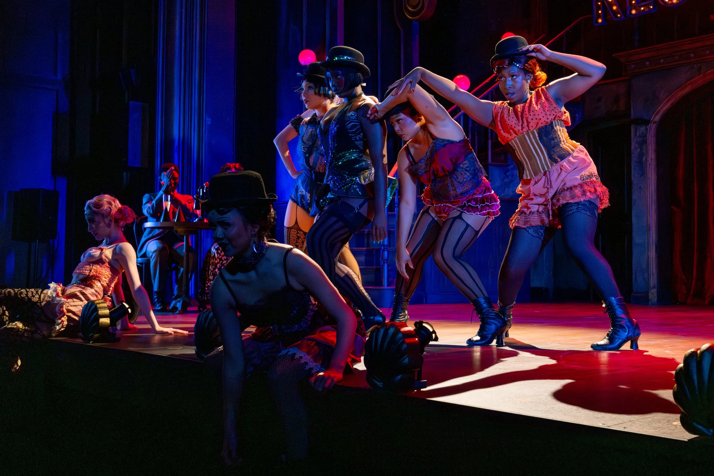A group of performers dressed in colorful, vintage-style costumes, many wearing hats, on a stage with dramatic lighting, some sitting and others standing, during a theatrical performance.
