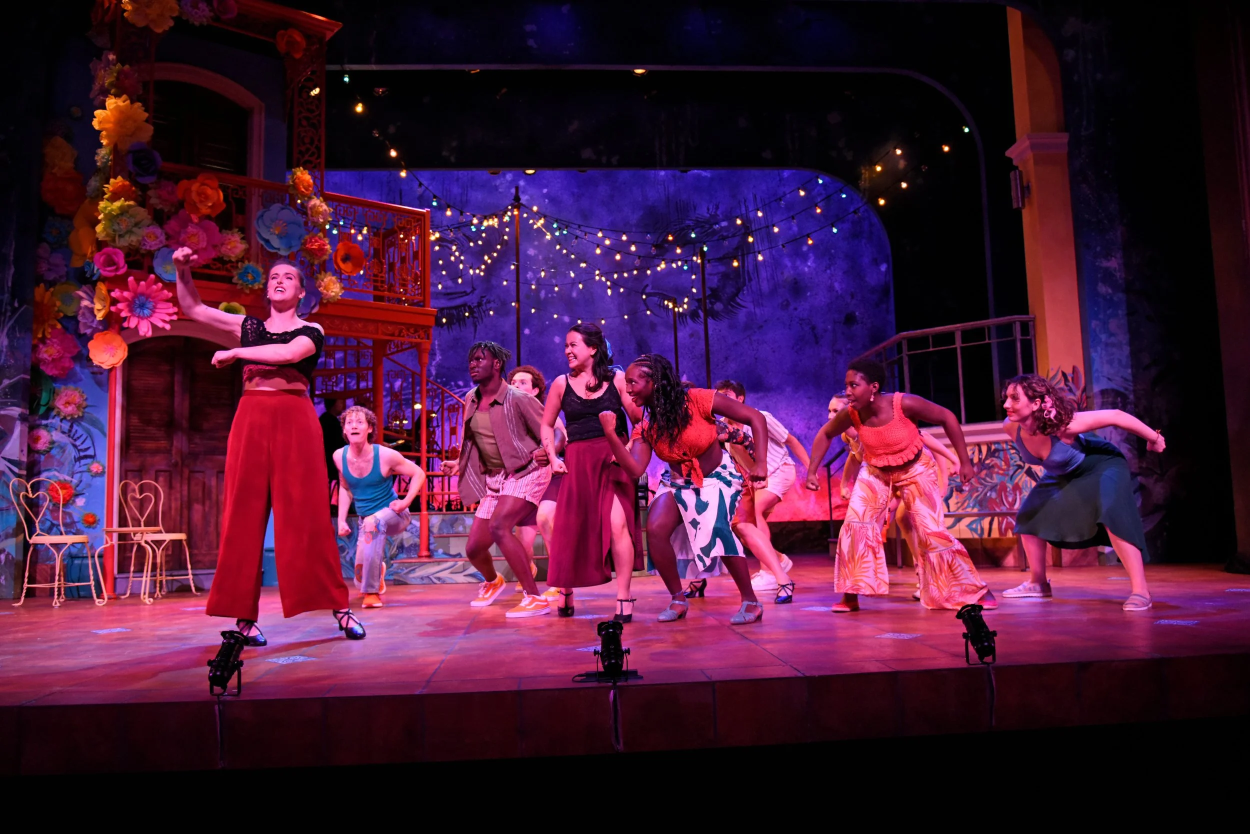 Theater stage with diverse group of performers dancing and singing, decorated with colorful flowers, string lights, and vibrant background.