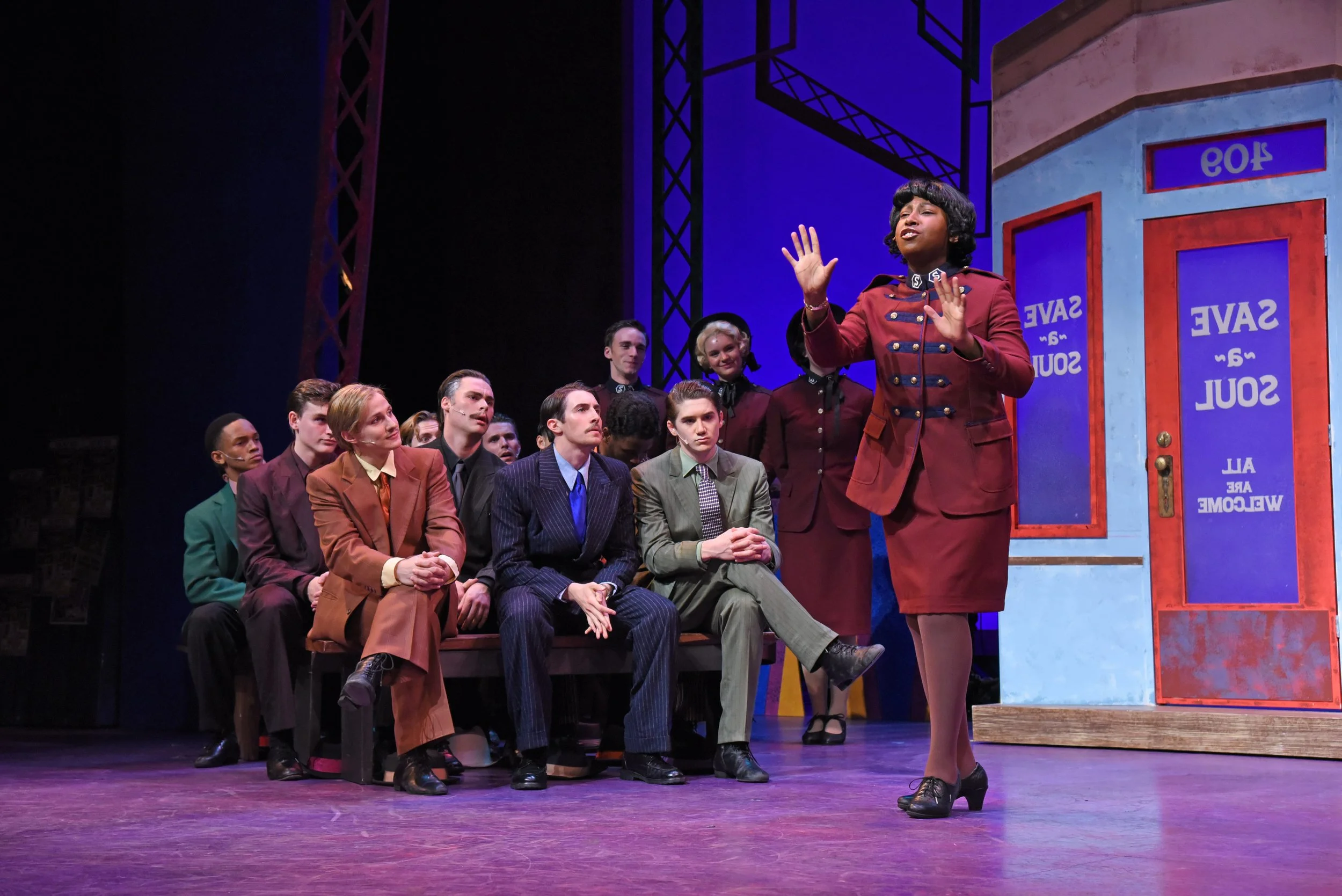 A woman in a red blazer and skirt speaking on stage in front of seated and standing audience members in a theater production. The scene appears to take place in a subway station or similar setting with blue and red painted doors in the background.