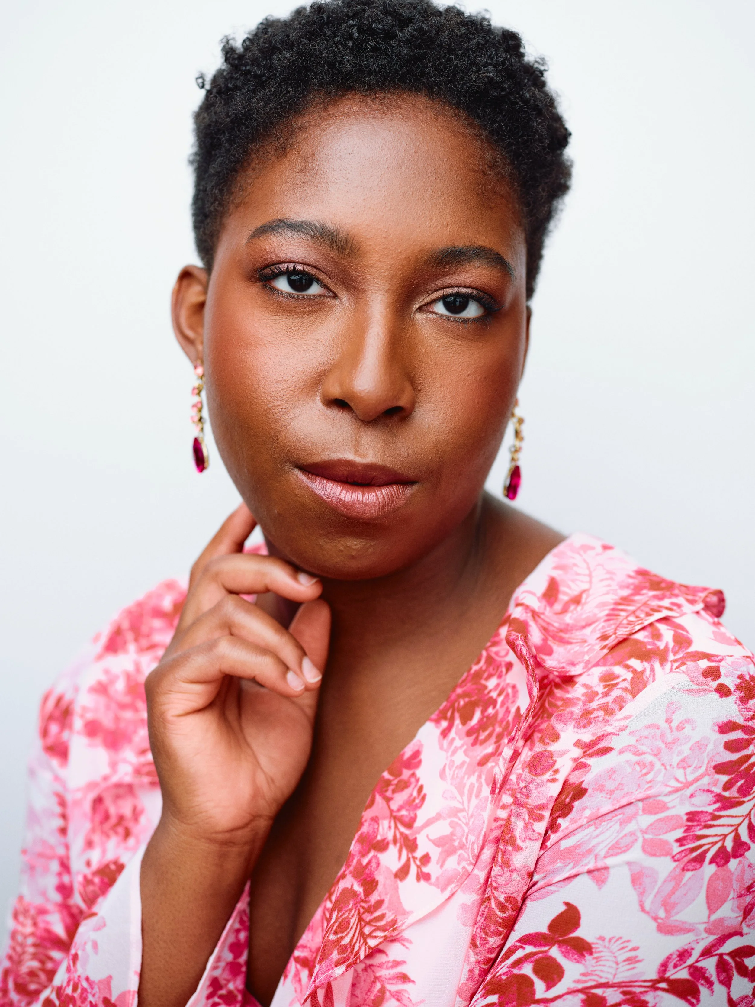 A woman with dark curly hair and brown skin poses with her hand gently touching her chin. She is wearing pink and white floral clothing and pink earrings, looking directly at the camera against a plain white background.