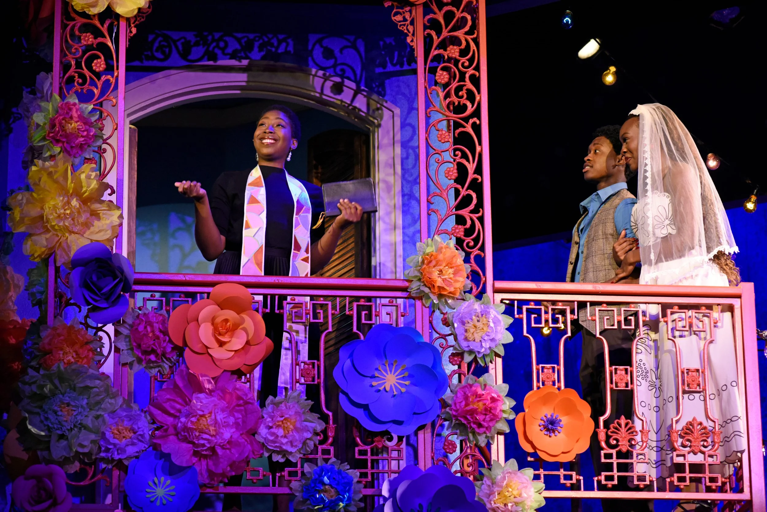 A theatrical scene with three actors on a decorated balcony. The woman at the center is speaking, holding a book, and is smiling. The other two actors, a man and a woman, are looking at her, dressed in costumes suggesting a wedding scene. The balcony is adorned with colorful paper flowers and intricate pink metalwork.