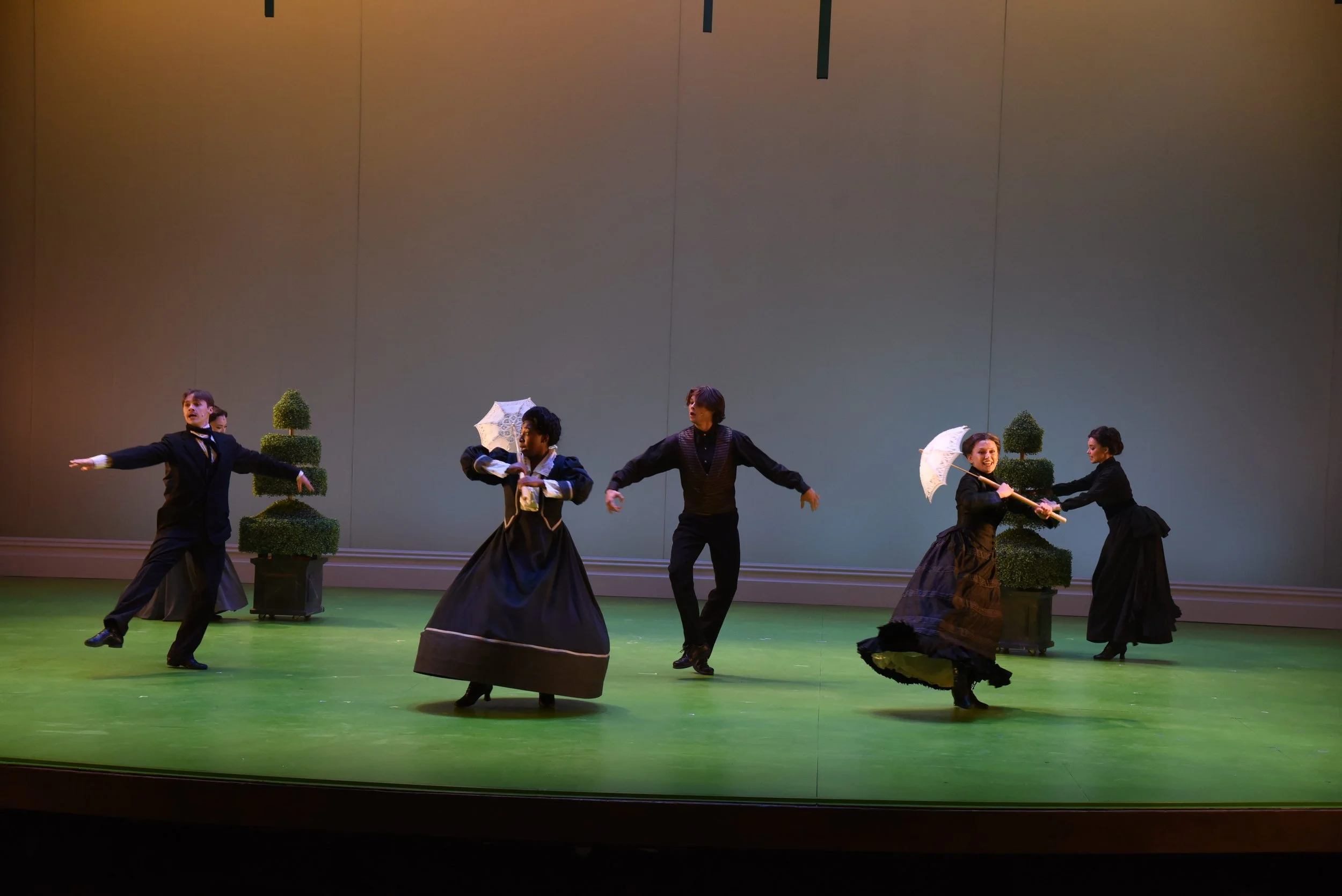 Theater scene with five performers in Victorian costumes, some holding umbrellas, on a stage with green floor and potted topiary.