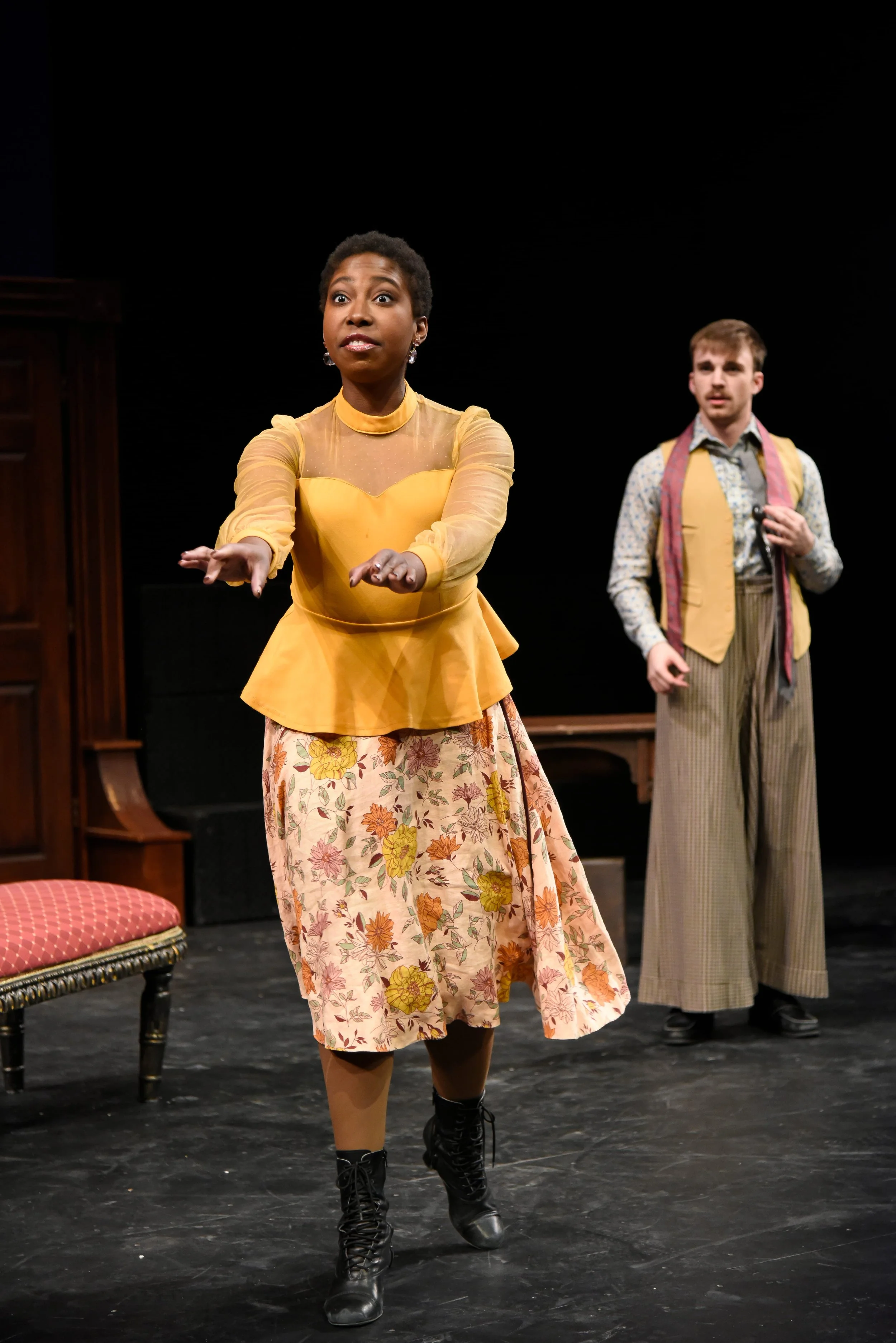 Two actors on stage, a woman in a yellow top and floral skirt in the foreground, and a man in patterned trousers and a vest in the background, performing in a play.