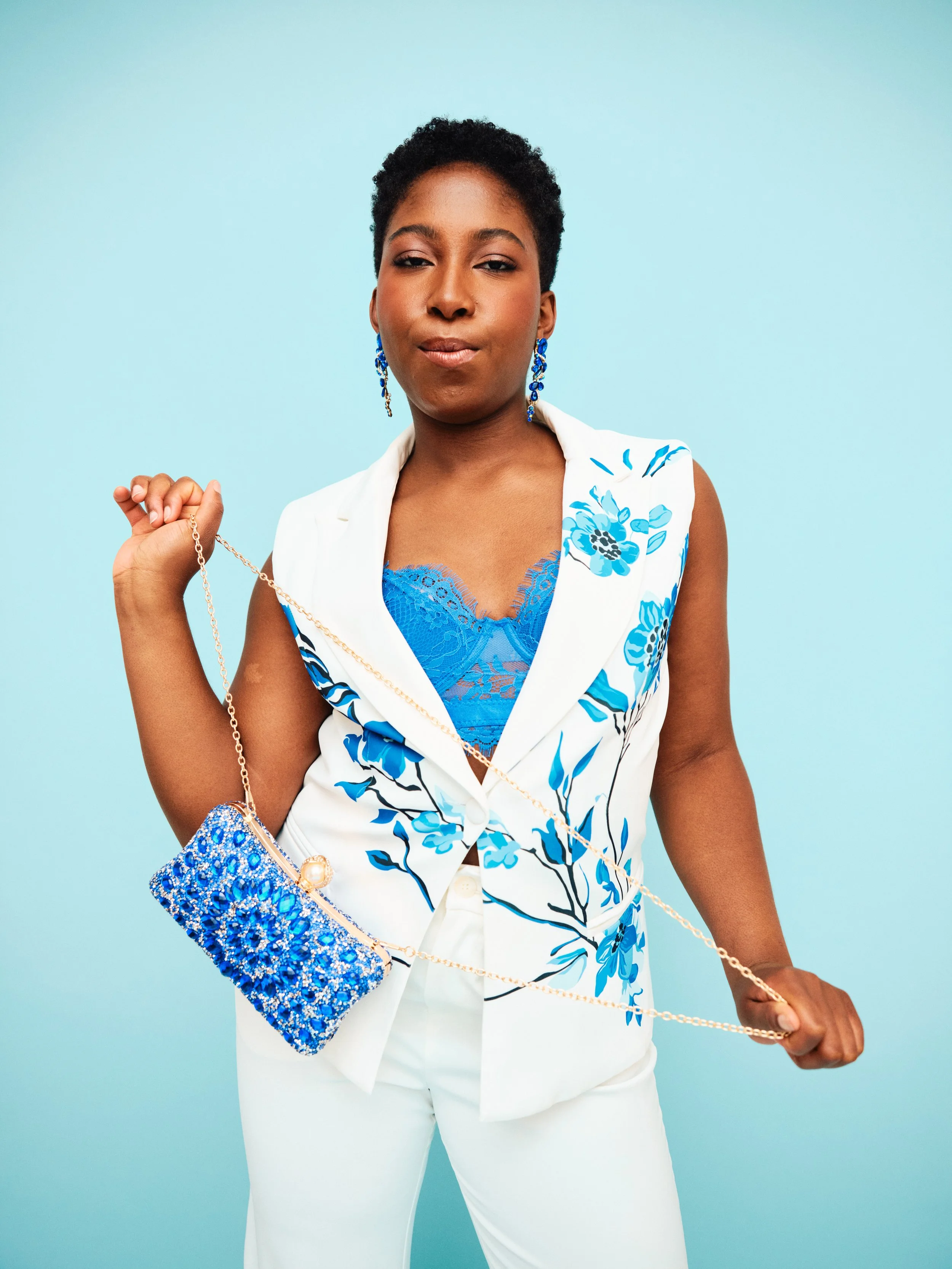 A confident woman with short curly hair wearing a white and blue floral sleeveless blazer, blue lace top, white pants, and blue jewelry, holding a blue beaded purse with a gold chain against a light blue background.