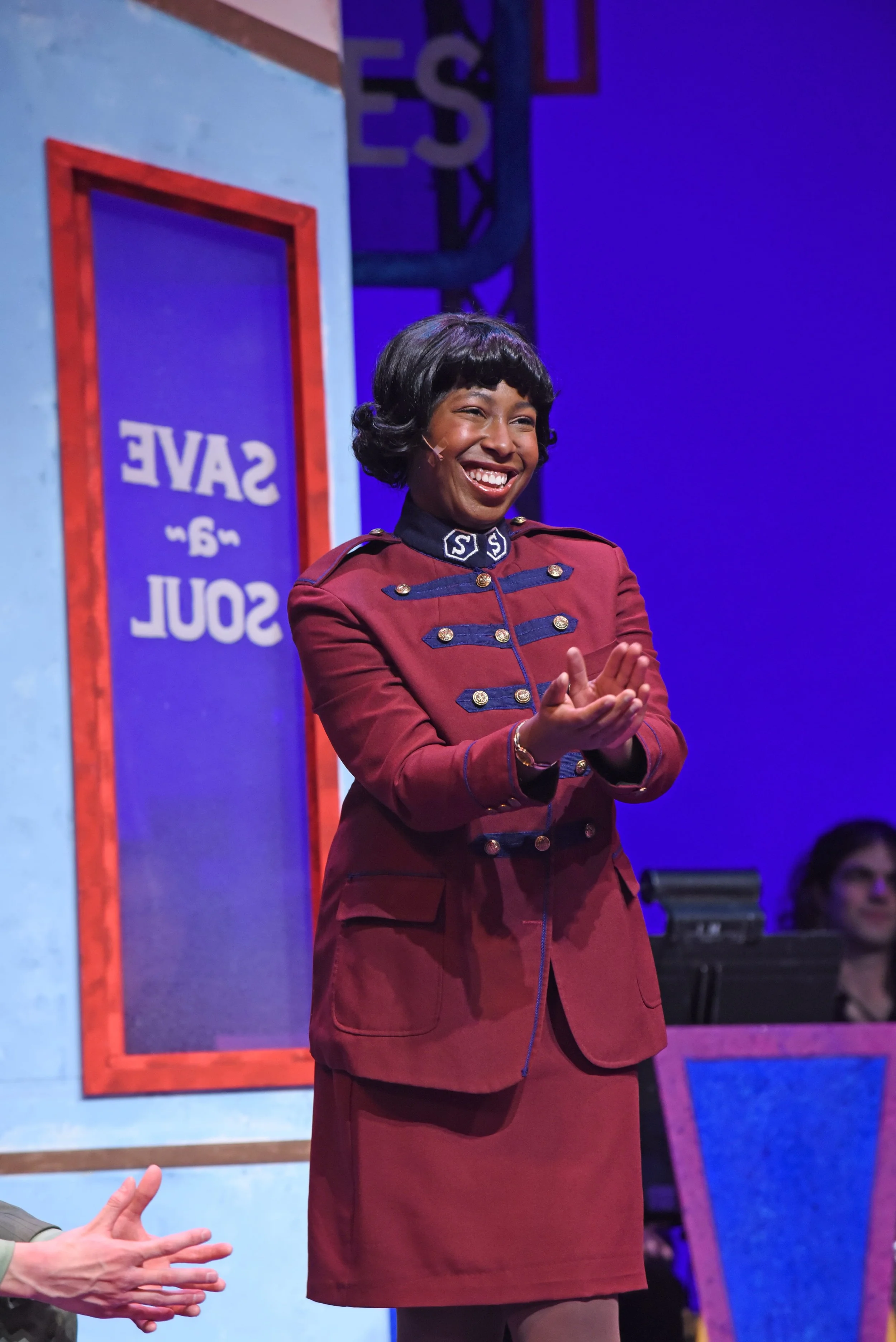 A woman in a red military-style uniform smiling and clapping on stage during a performance or presentation.