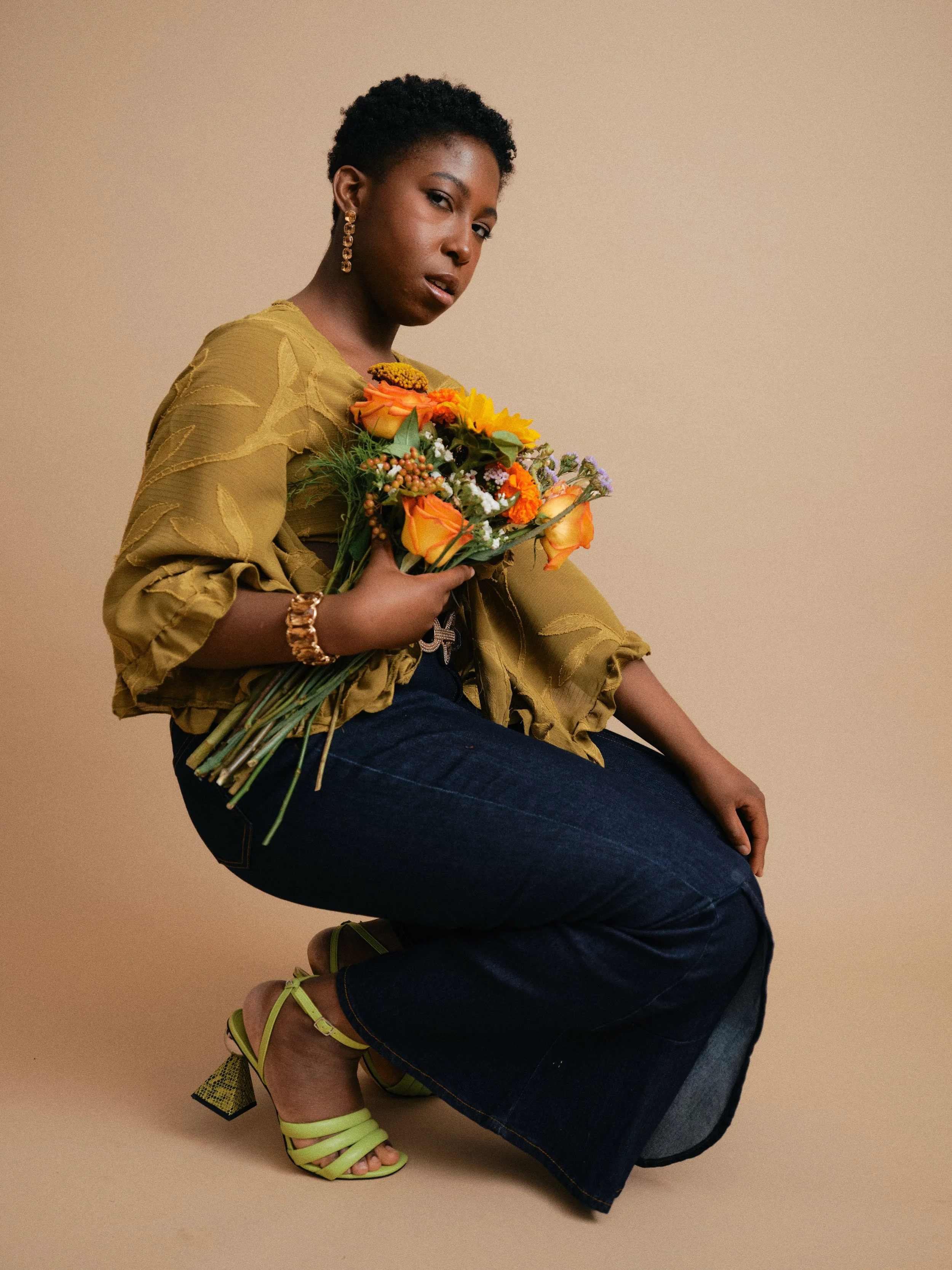 A woman with short curly hair, wearing a yellow blouse, dark jeans, and lime green high heel sandals, is holding a bouquet of colorful flowers and posing against a beige background.