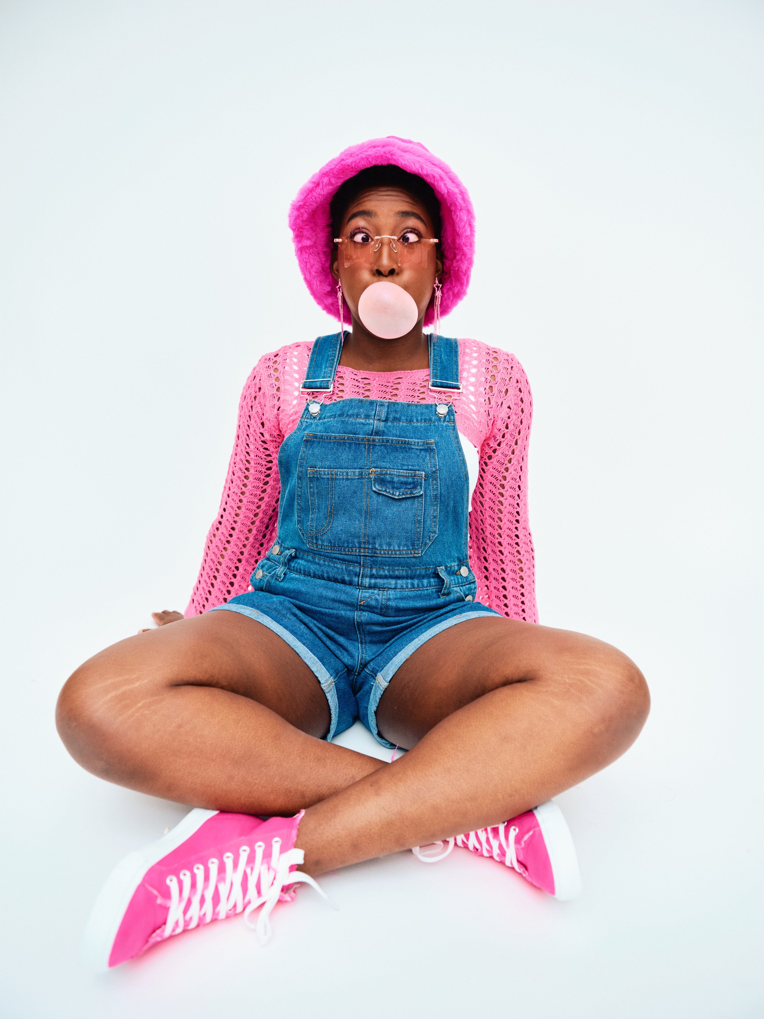 A young woman sitting cross-legged on the floor blowing a bubble with bubblegum, wearing pink sneakers, denim overalls, a pink lace top, a pink fuzzy hat, and glasses, against a plain white background.