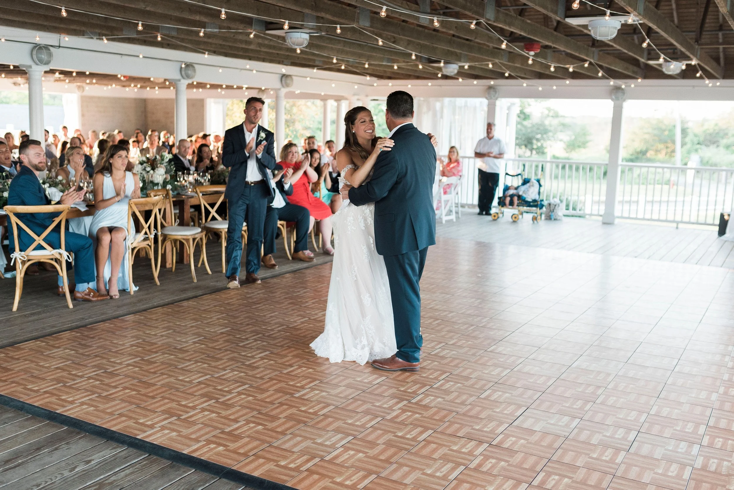 Bride and groom dancing at their wedding reception, surrounded by seated and standing guests, under string lights in a decorated indoor/outdoor venue.