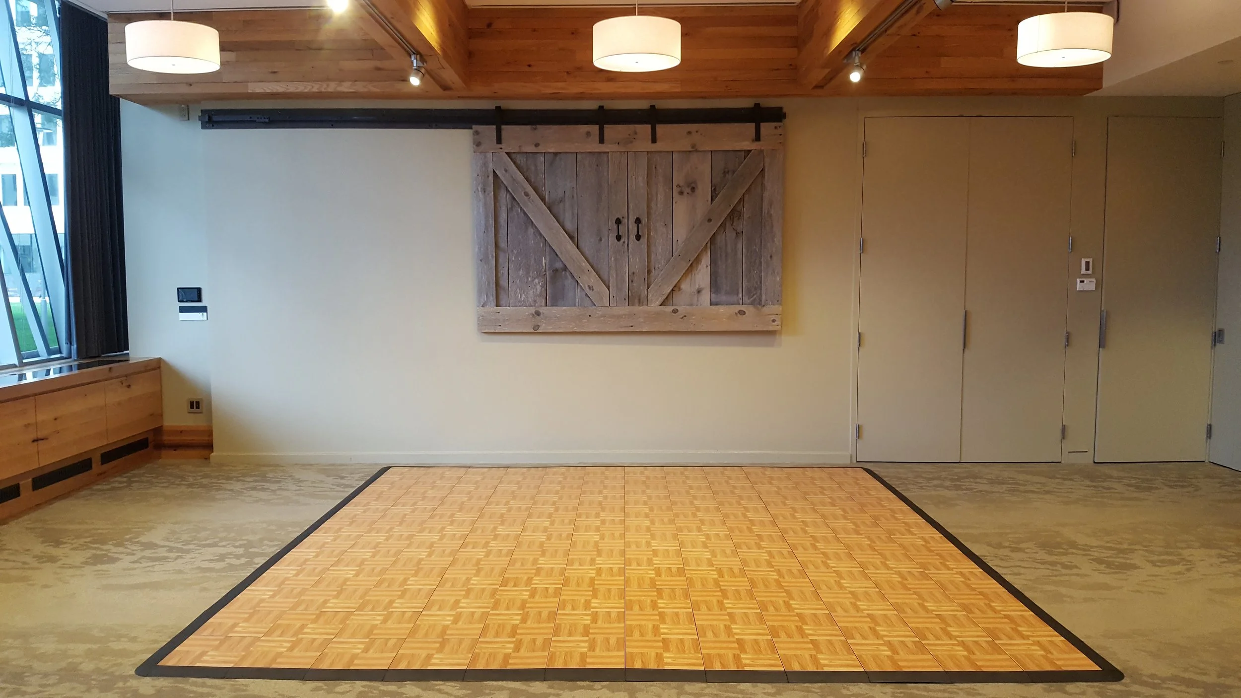 Empty room with wooden ceiling and beige walls, featuring a wooden sliding barn door on the back wall, a small indoor dance floor in the center, a large window on the left side, and multiple light fixtures on the ceiling.