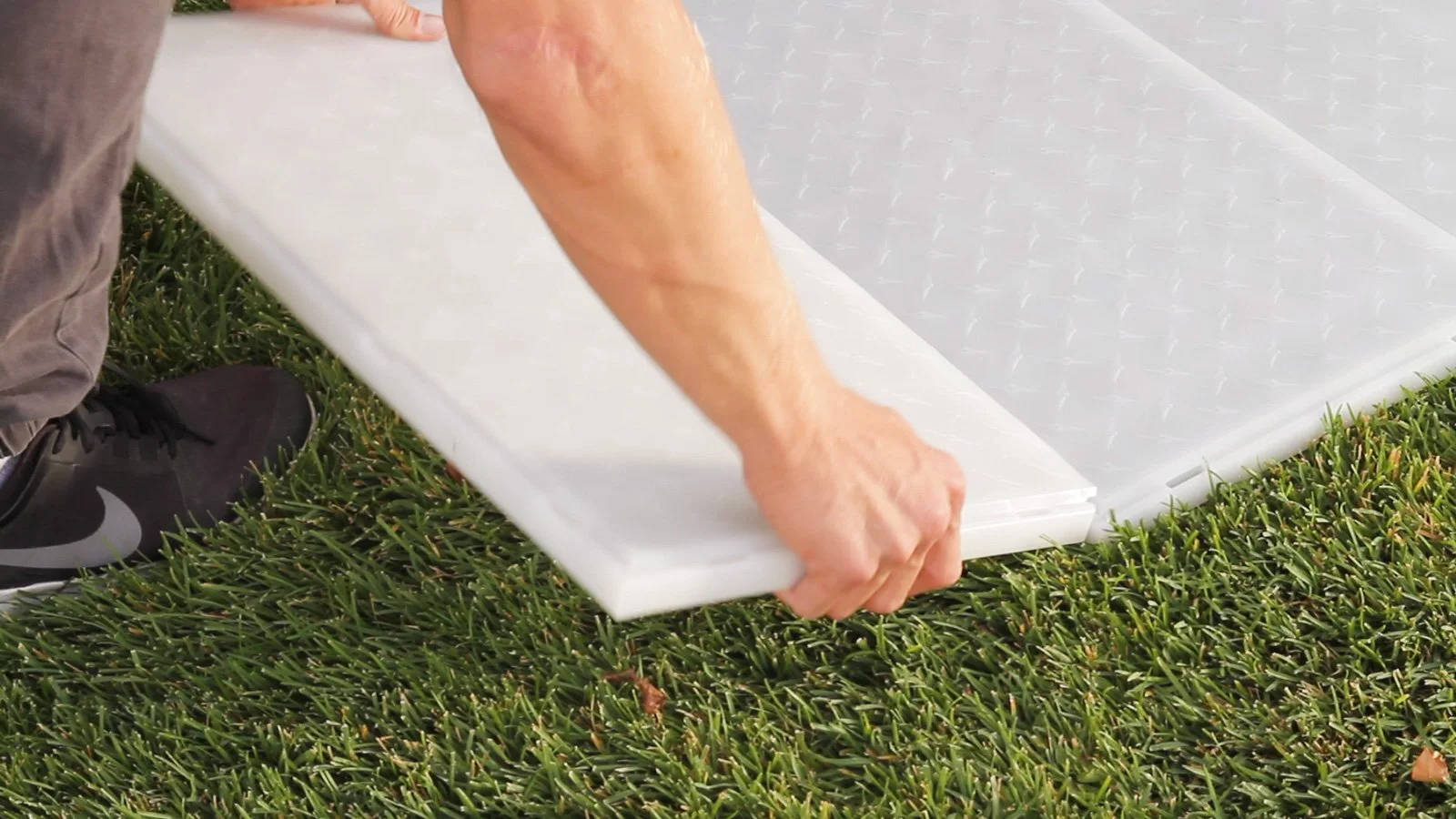 A person installing a white plastic roof panel on a grassy area, wearing black Nike sneakers.