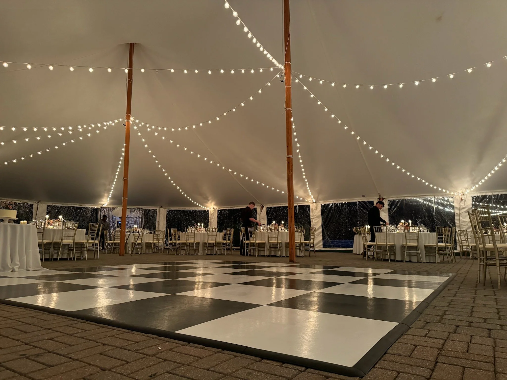 Decorative event tent with string lights, round dining tables with white tablecloths, and gold chairs, set for a celebration, with a black and white checkered dance floor in the foreground.