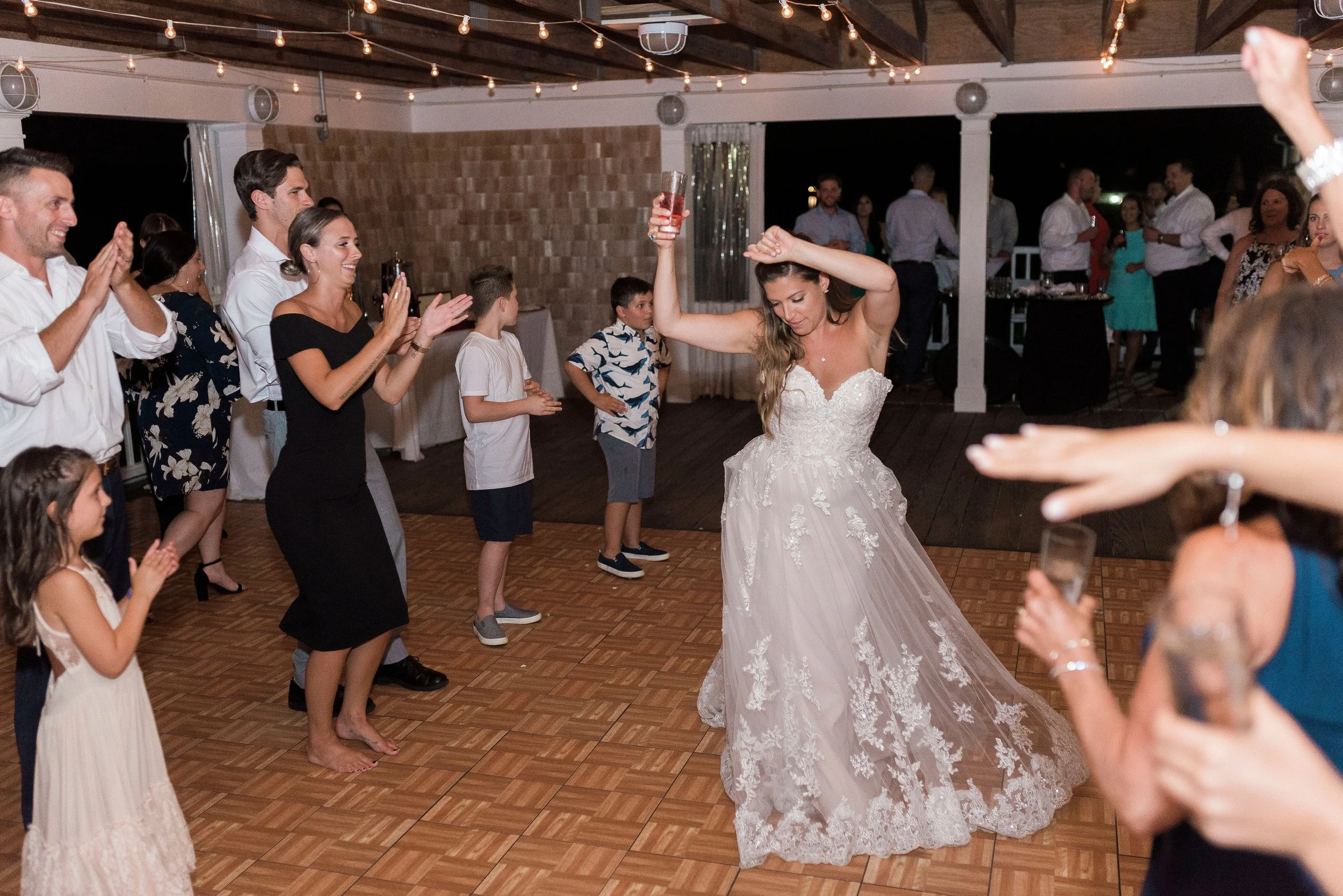 A bride in a white wedding dress dancing with her arm raised at a wedding reception, surrounded by guests clapping and smiling on a wooden dance floor under string lights.