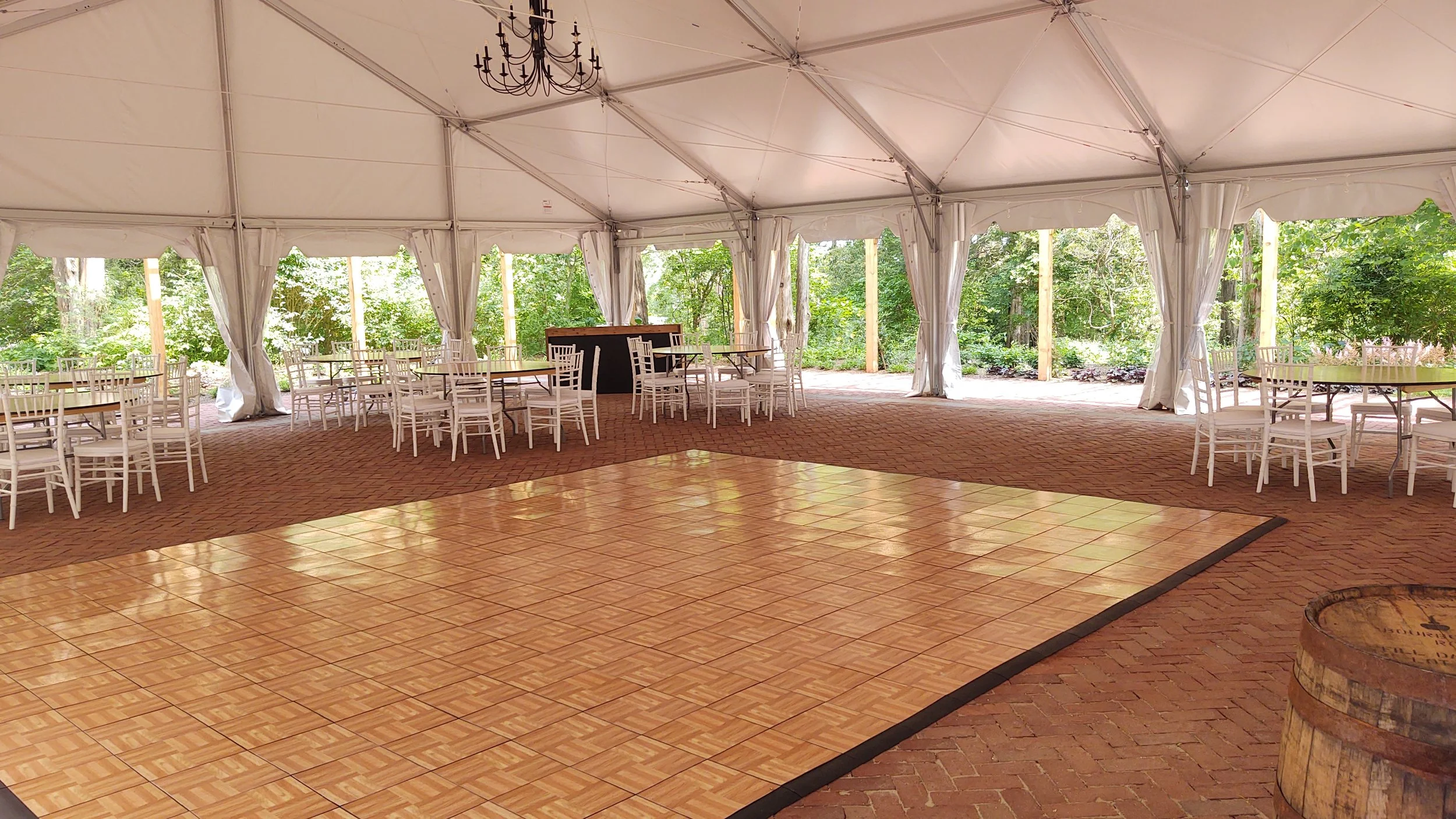 Empty event tent with a large dance floor, white chairs, and tables, surrounded by trees and greenery.